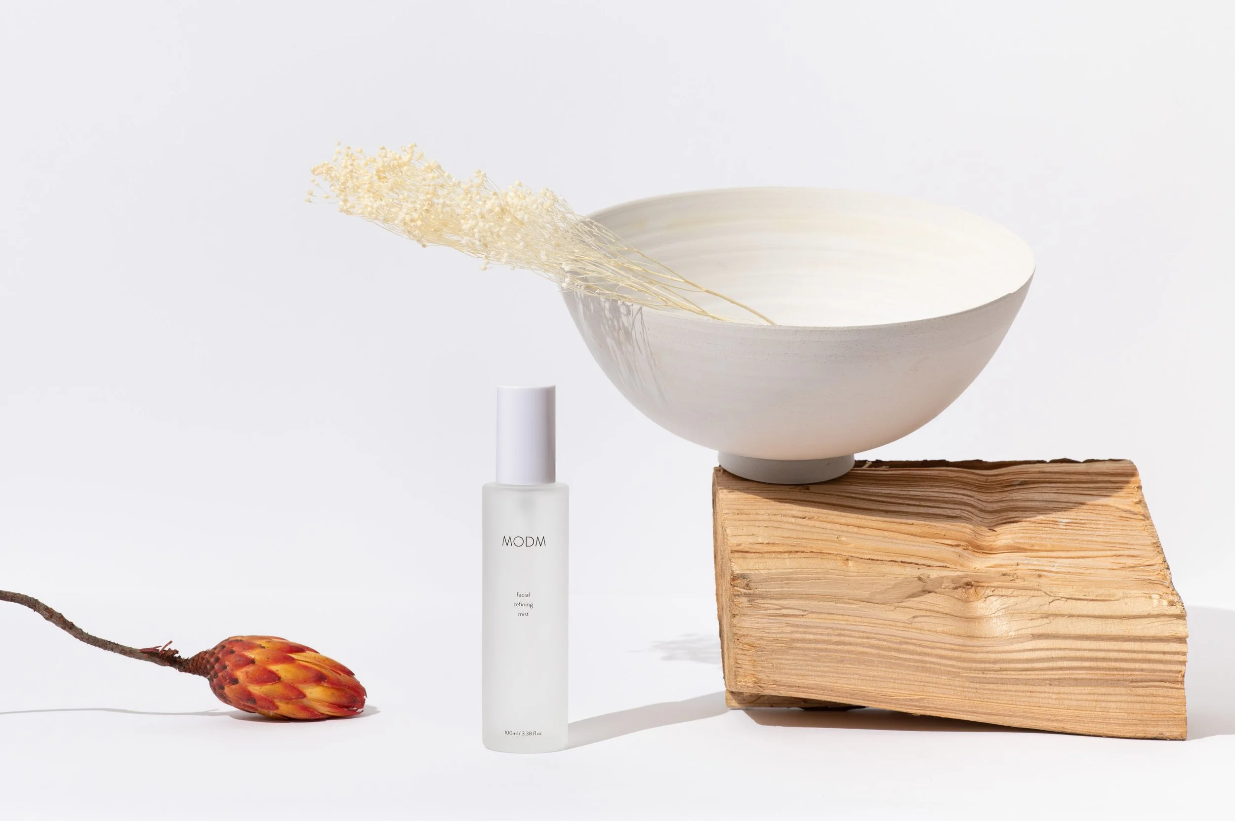 Minimalist still life with a white ceramic bowl on a wooden block, a white mist spray bottle labeled MODM, a dried flower, and a small orange and red fruit on a plain white background.