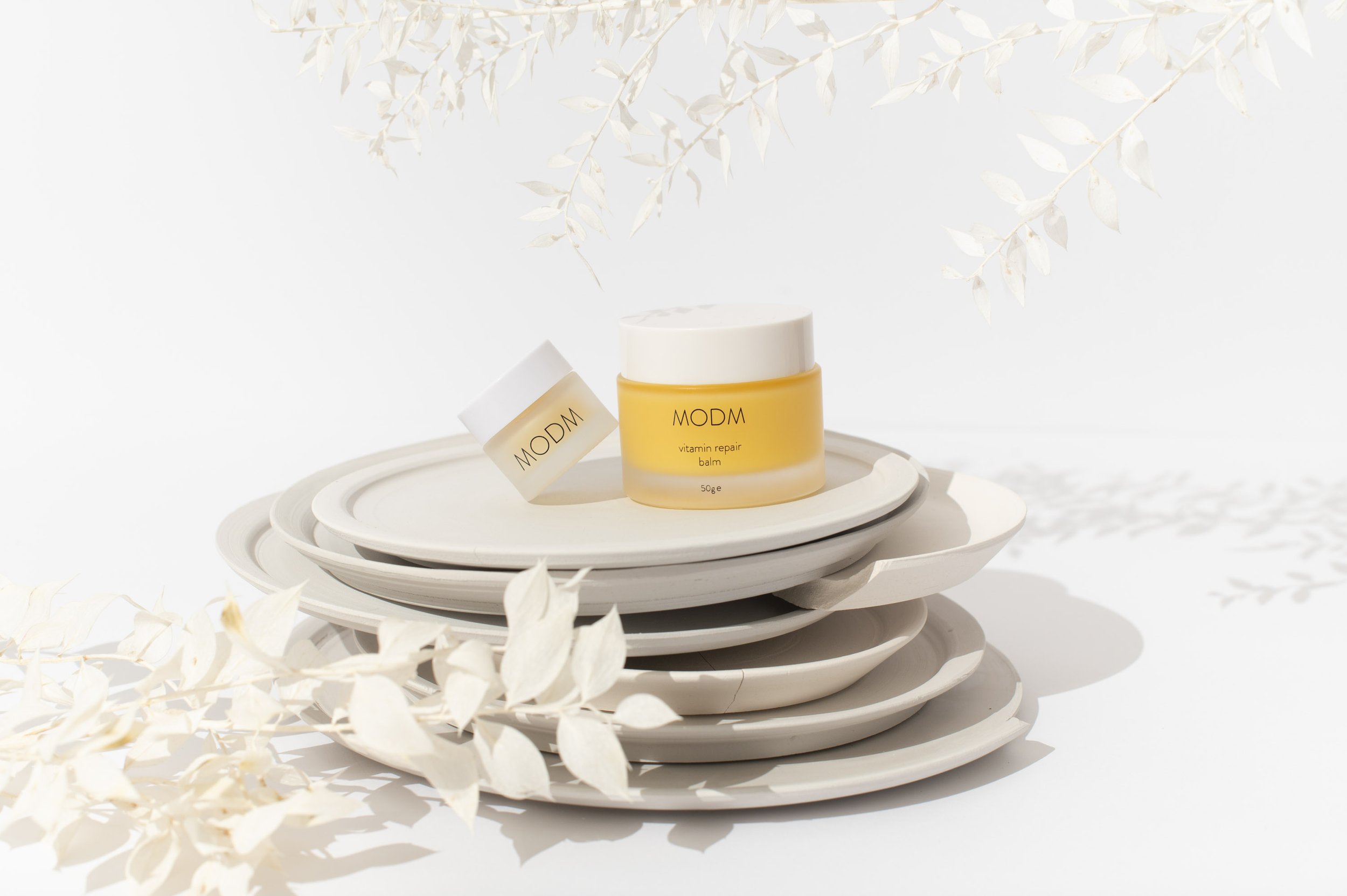 Stack of white plates with two jars of MODM vitamin repair balm, set against a white background with white leaves.