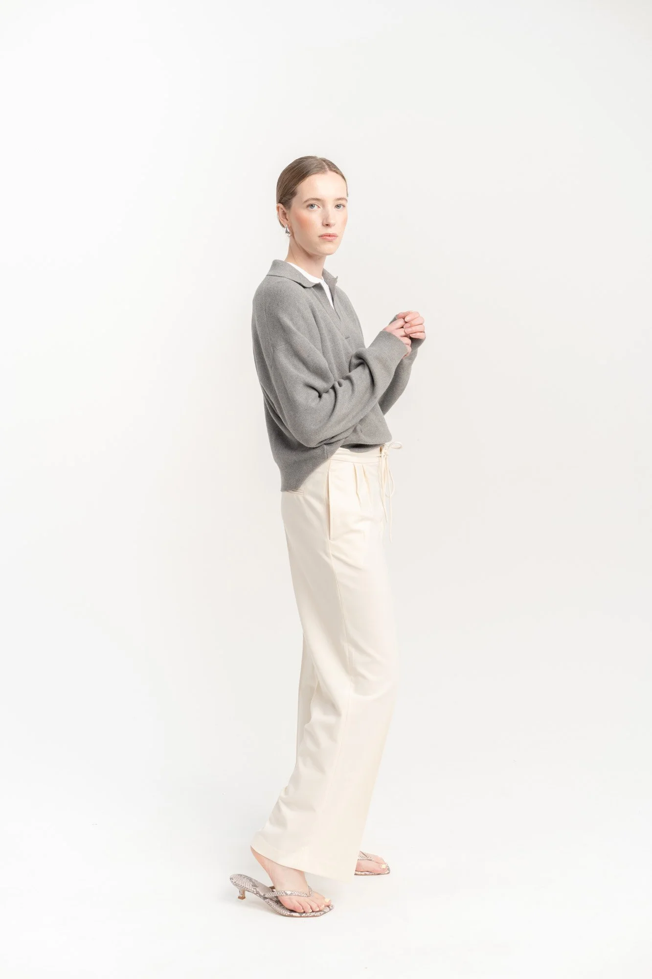 A woman with brown hair tied back, wearing a gray sweater, white pants, and open-toed heels, standing with her hands clasped in front of her against a plain white background.