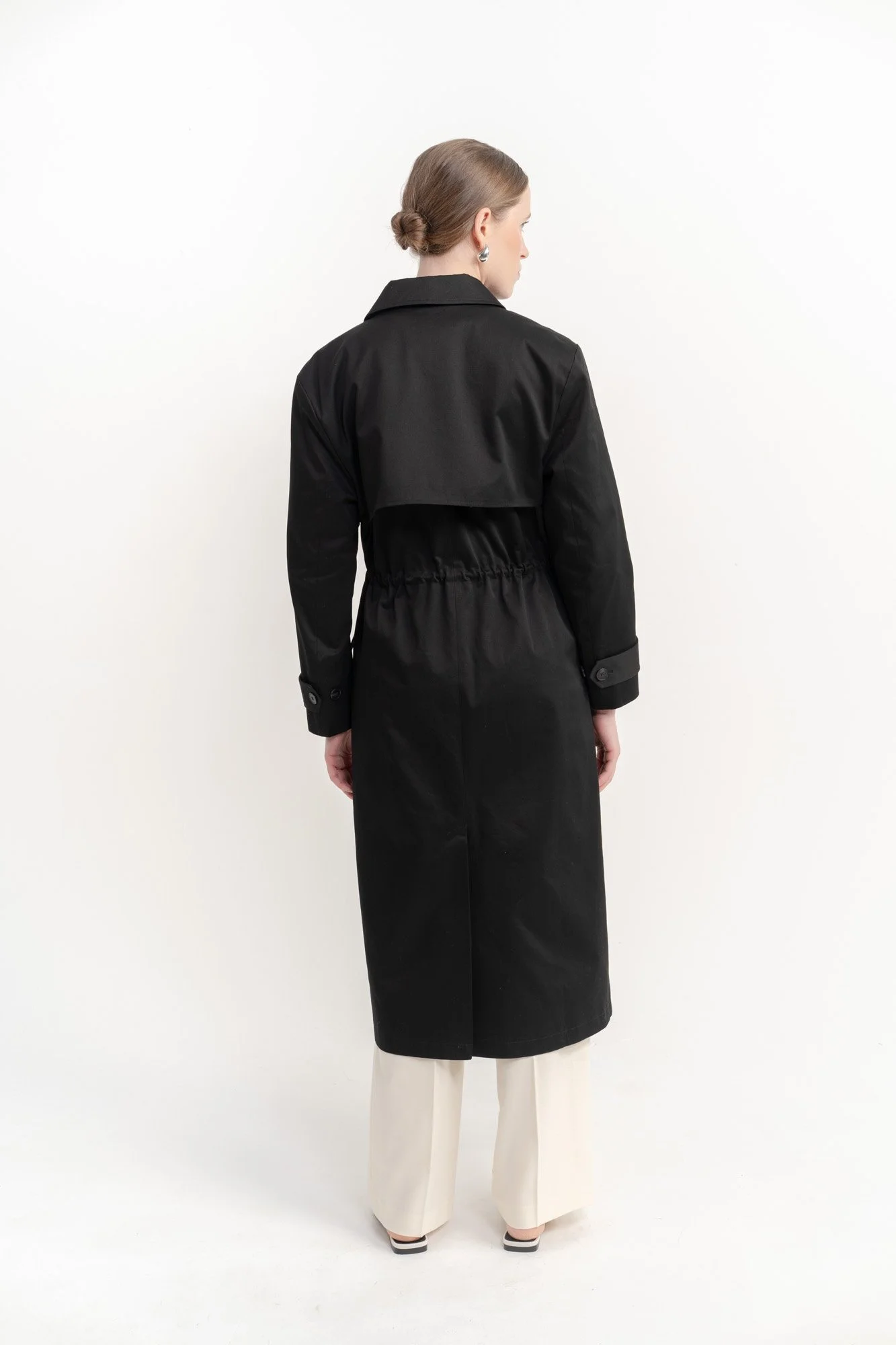 Back view of a woman wearing a long black trench coat and cream-colored wide-leg pants, standing against a plain white background.