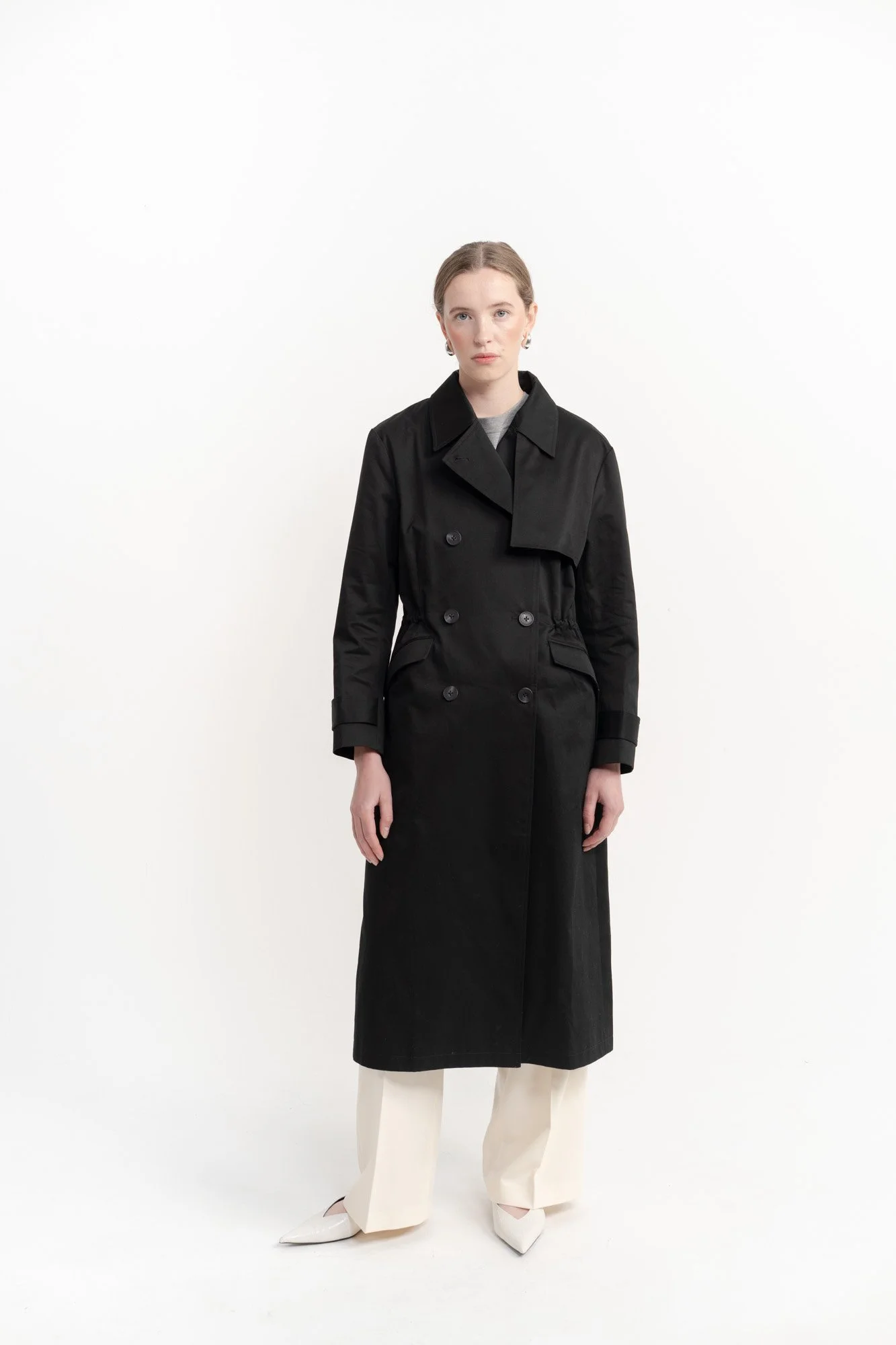 A woman wearing a black trench coat, light-colored wide-leg pants, and white shoes, standing against a plain white background.