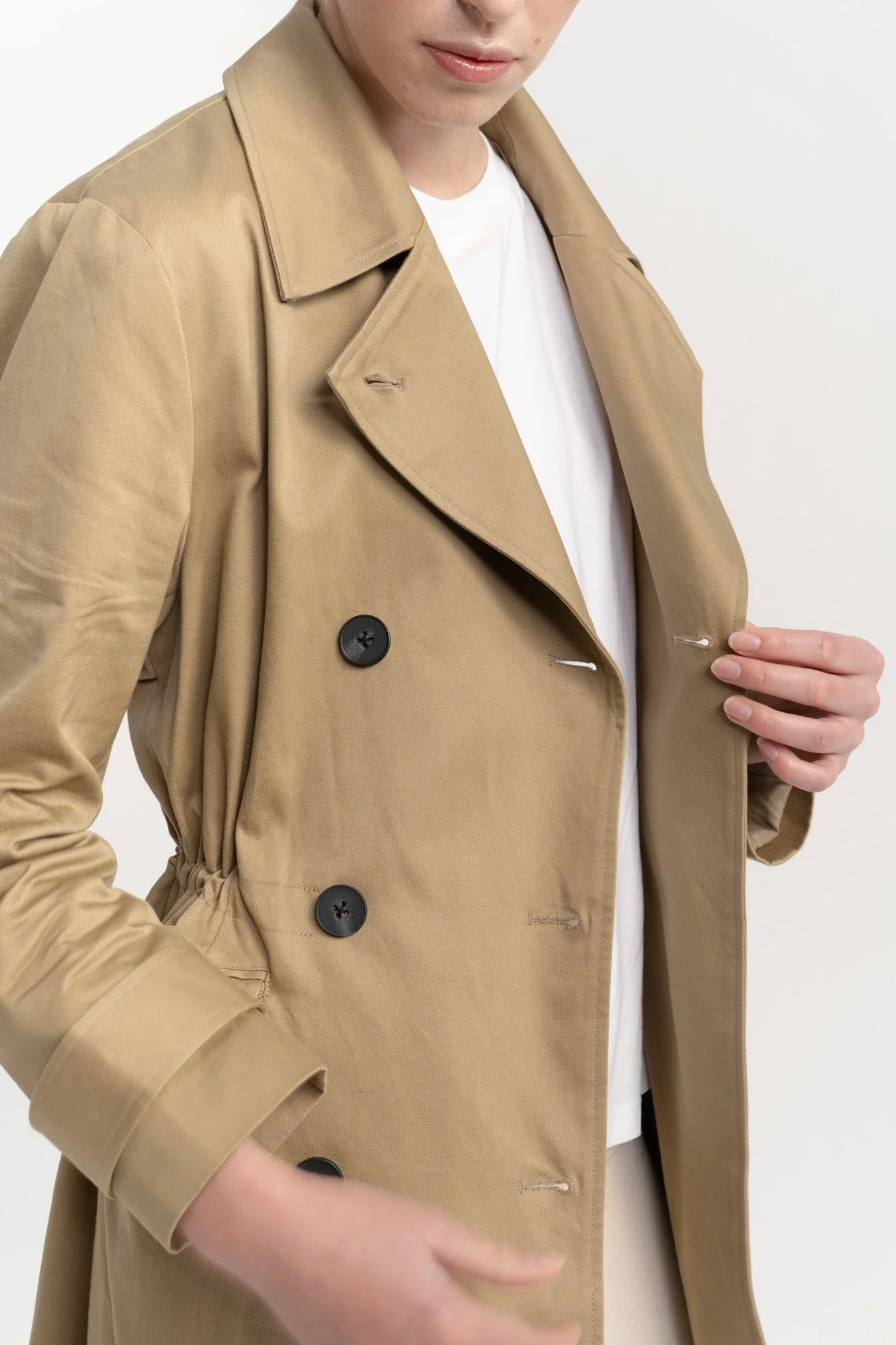 Close-up of a woman wearing a beige trench coat over a white shirt, with her hand adjusting the coat.