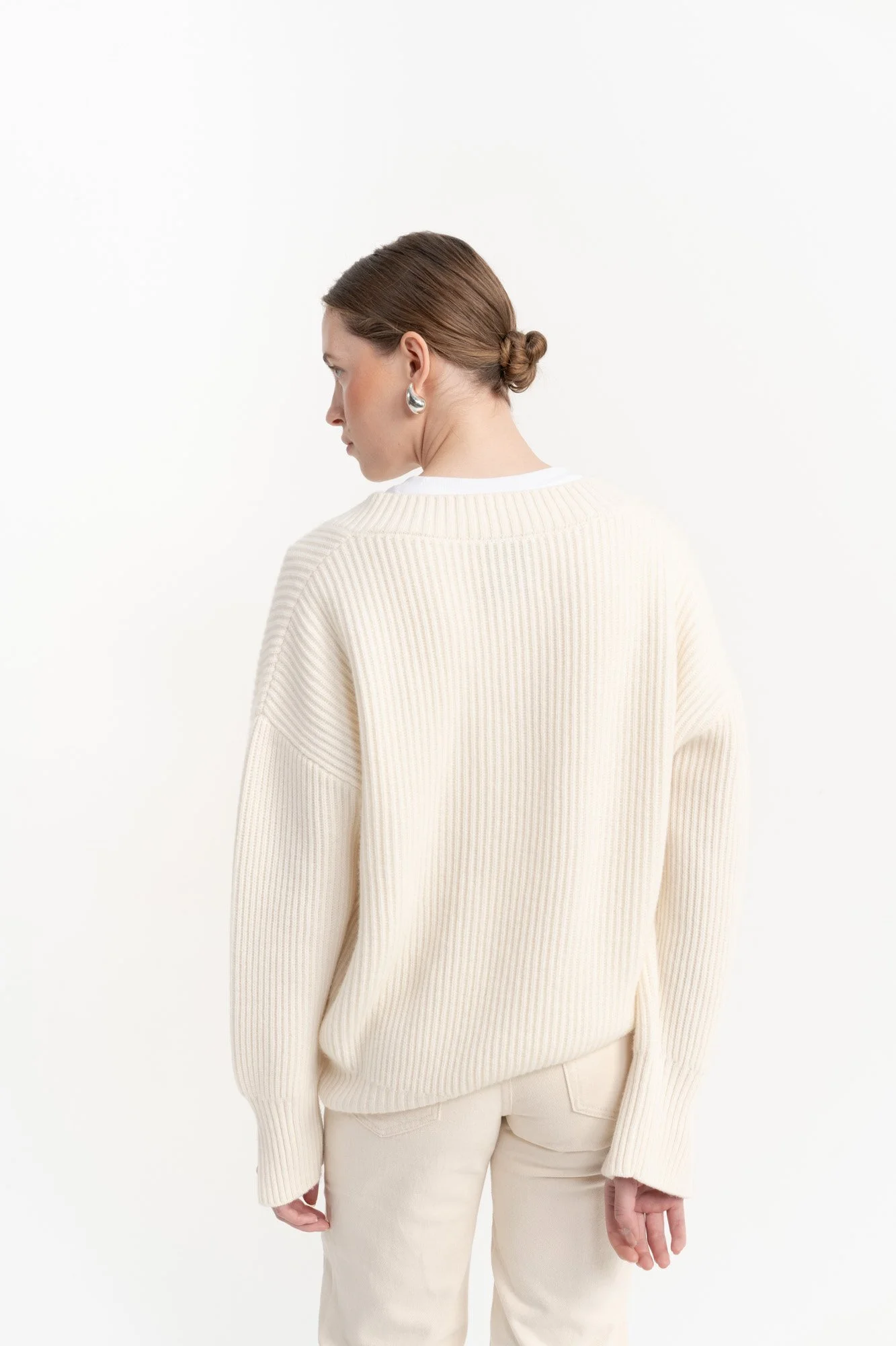 Back view of woman with brown hair in a bun wearing a cream oversized ribbed sweater and cream pants, standing against a white background.