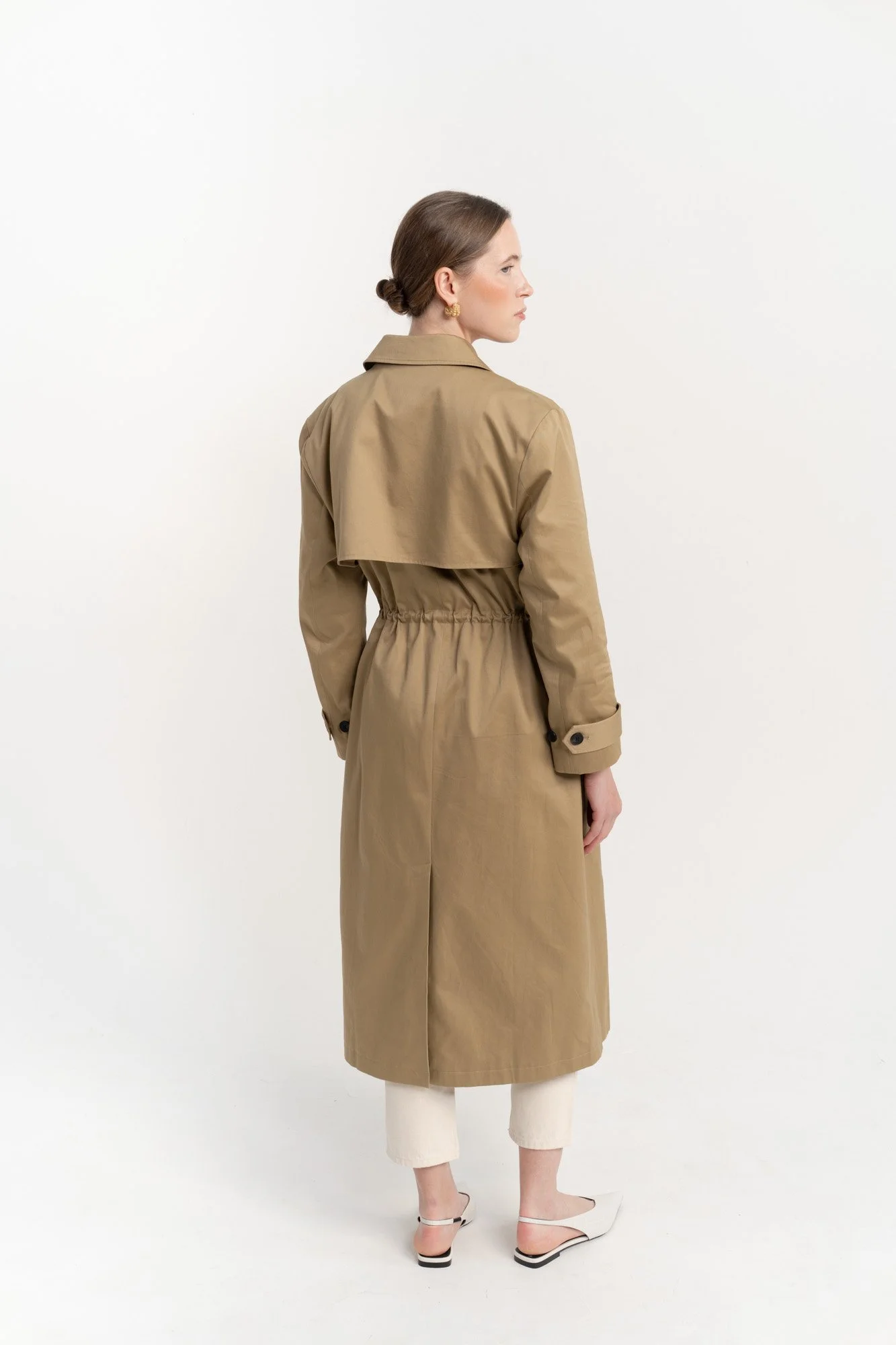 Back view of a woman in beige trench coat, white pants, and white heels standing against a white background.