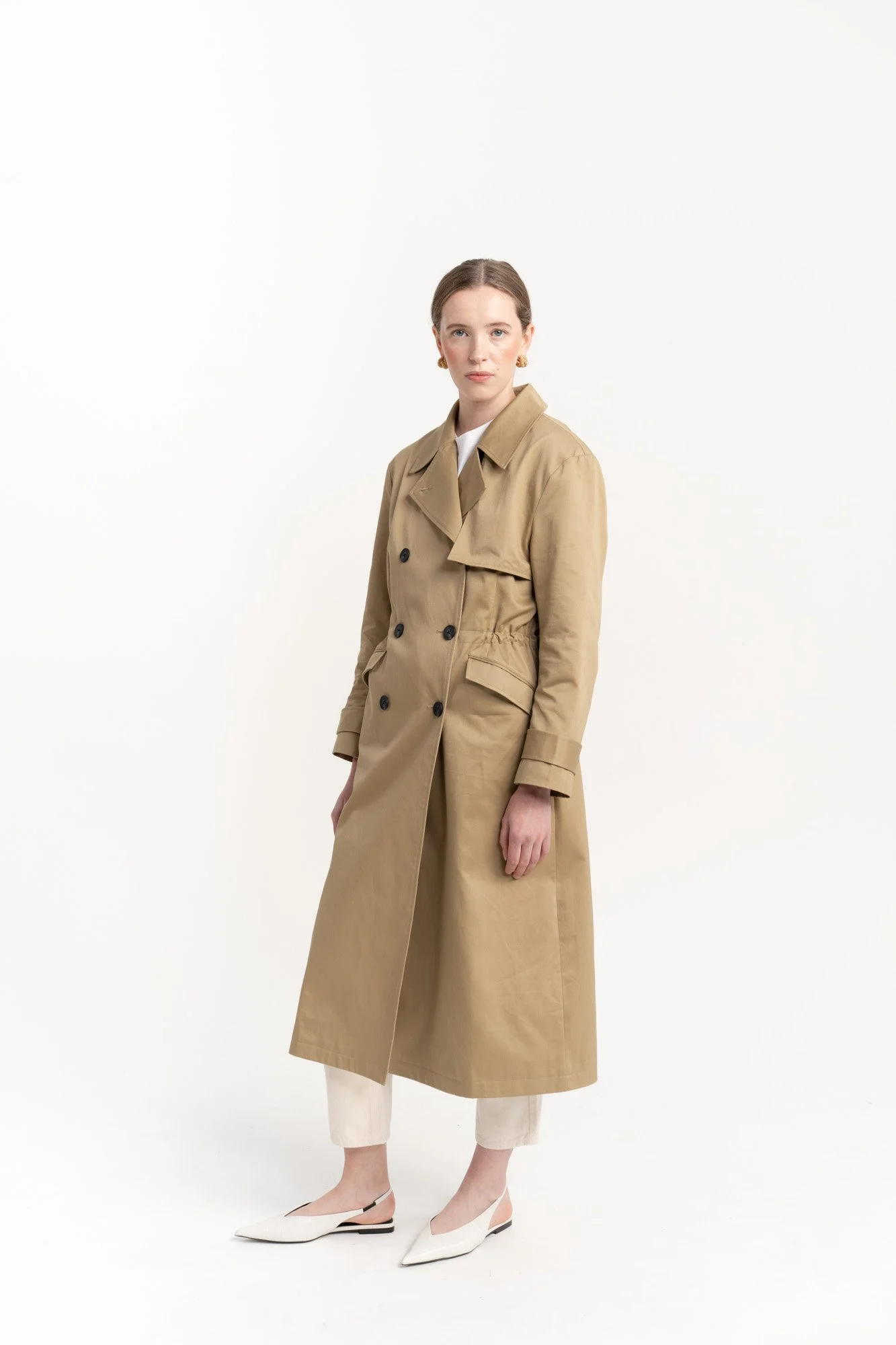 A woman wearing a tan trench coat, white pants, and white shoes standing against a white backdrop.