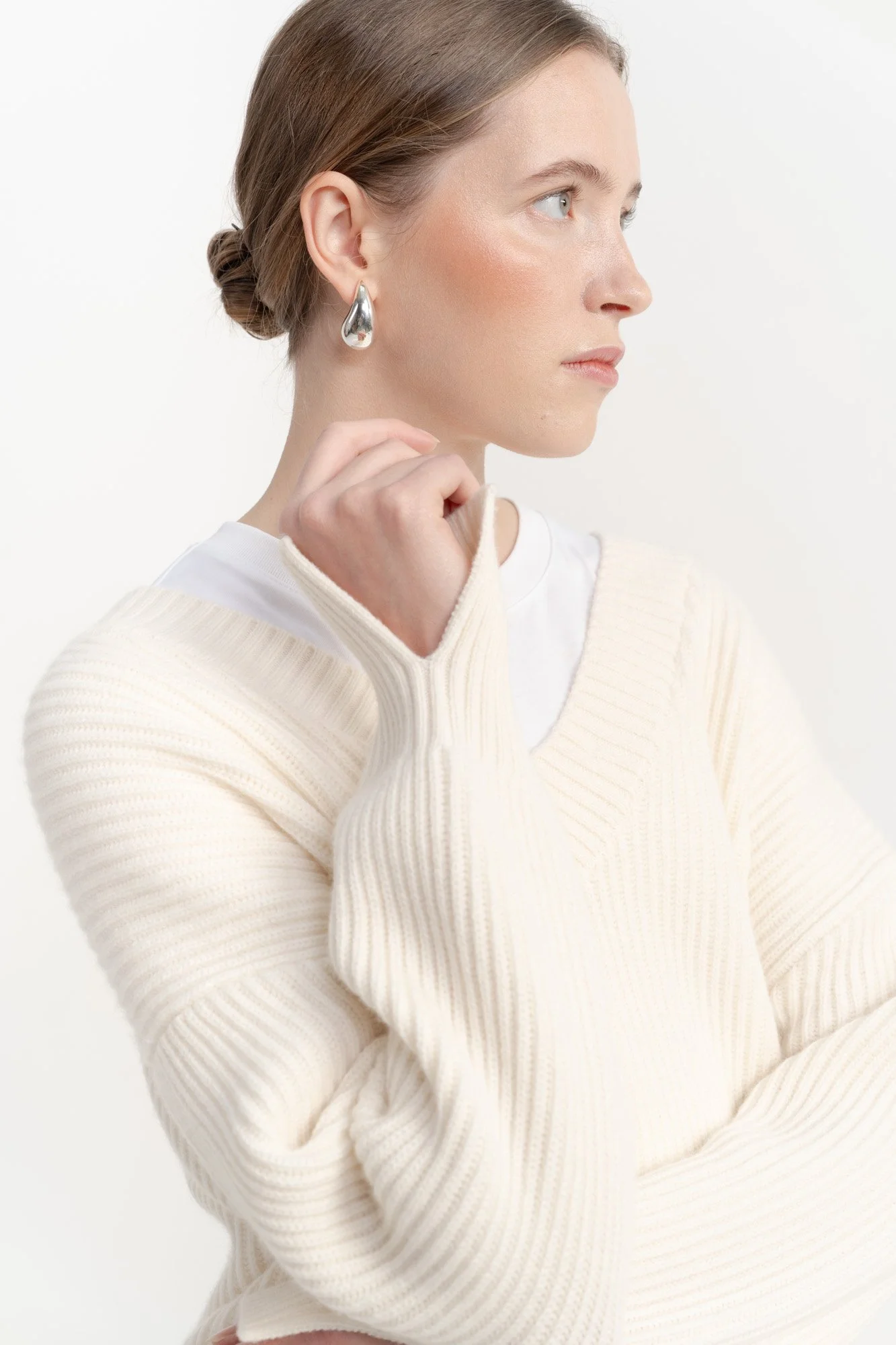 A woman with short brown hair styled in a low bun, wearing a white sweater over a white shirt, silver earrings, and looking to the right against a plain white background.
