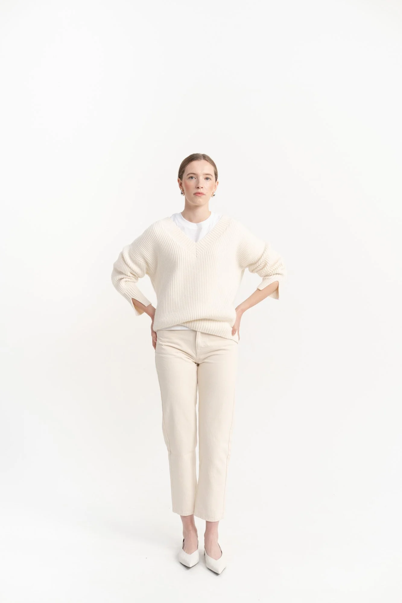 A young woman stands with her hands on her hips, wearing a cream-colored oversized sweater, cream pants, and white heels, against a plain white background.