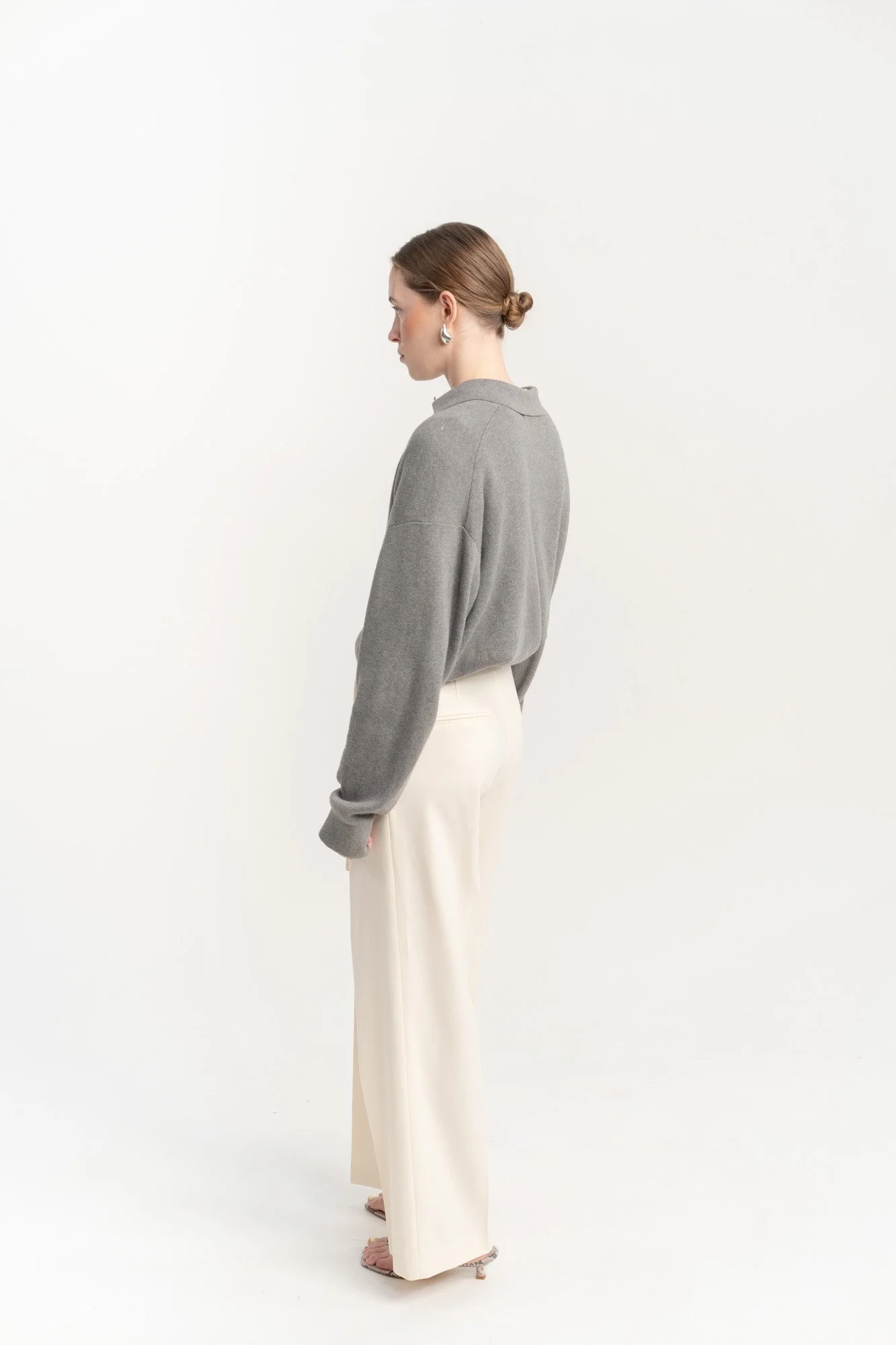 A woman with her hair in a bun, wearing a gray oversized sweater, cream-colored wide-leg pants, and open-toe heels, standing sideways against a plain white background.