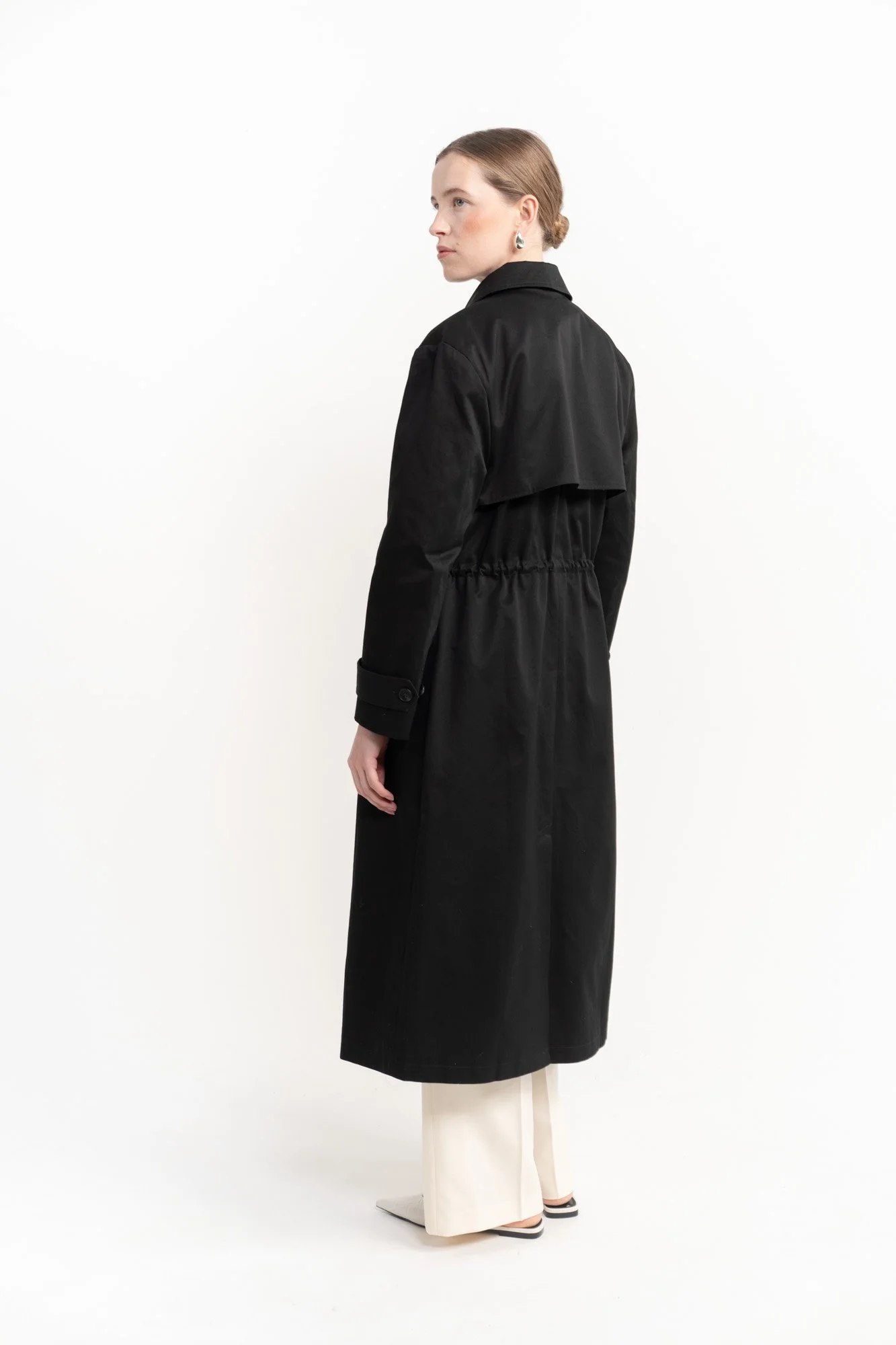 A woman wearing a long black raincoat, cream-colored pants, and pointed shoes, standing against a plain white background.