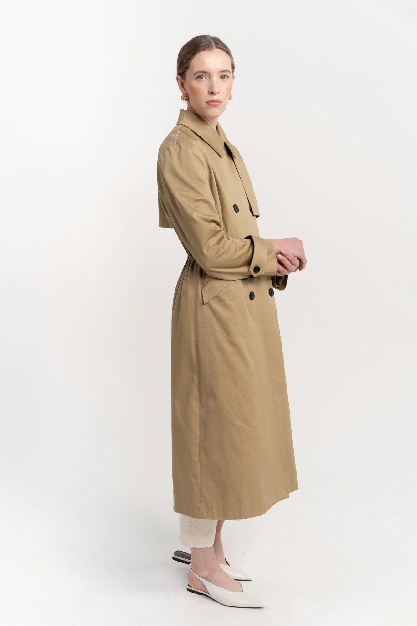 A woman wearing a tan trench coat, white pants, and beige heels, standing against a plain white background.