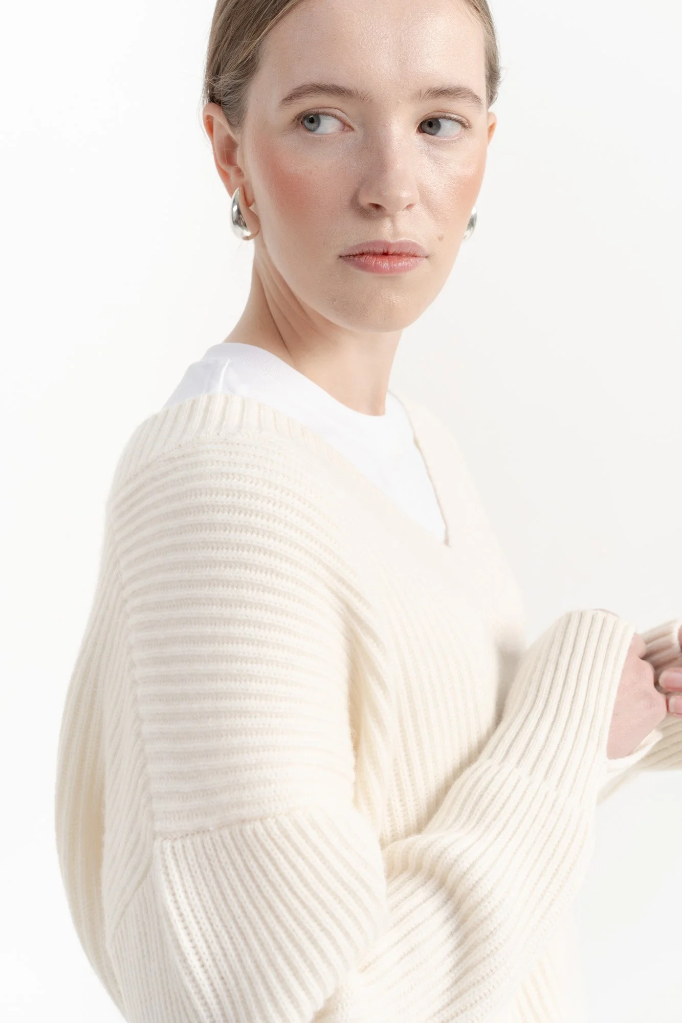 A woman with fair skin, light brown hair, and blue eyes looking to her right. She is wearing silver hoop earrings, a cream-colored ribbed sweater over a white shirt, and is holding her hands together.