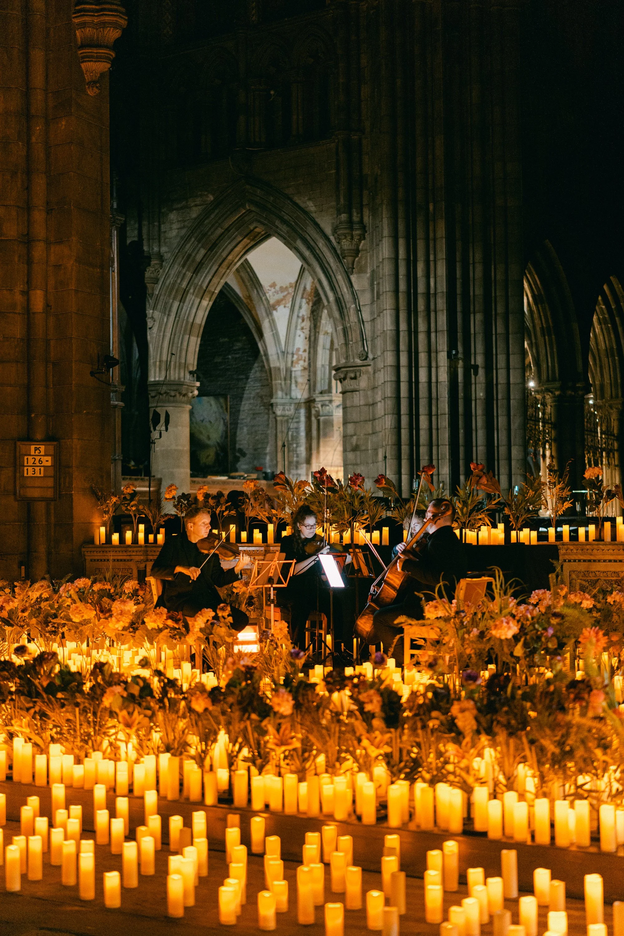 A musical performance in a church with three musicians playing string instruments, surrounded by candles and flowers, with gothic arches overhead.