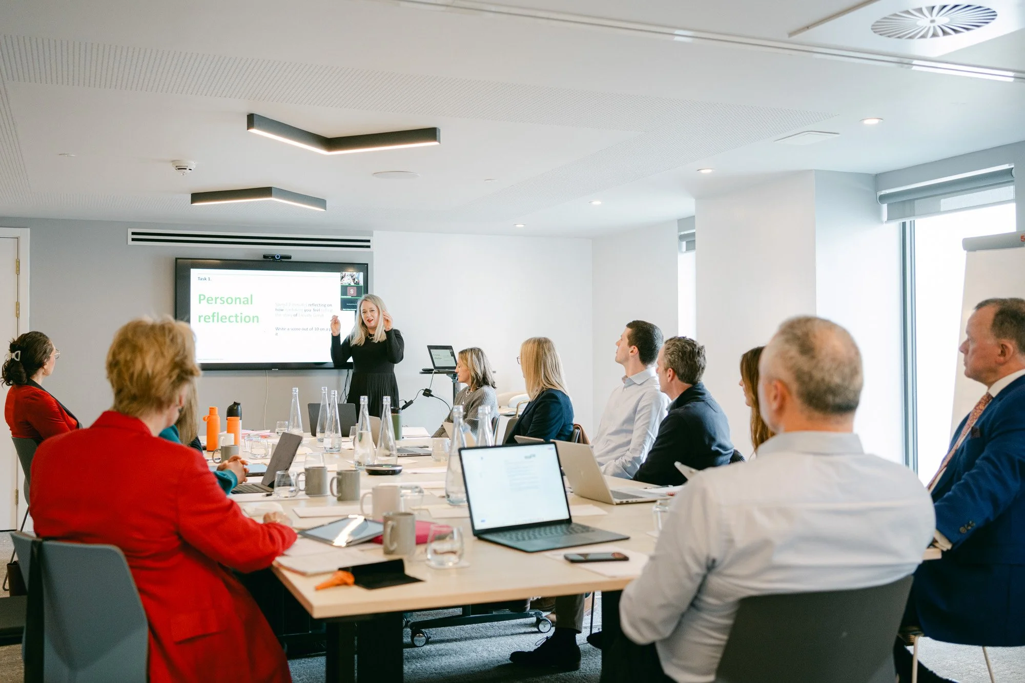 Business meeting in a conference room with a woman presenting to a group of seated people using a screen that reads 'Personal reflection'.