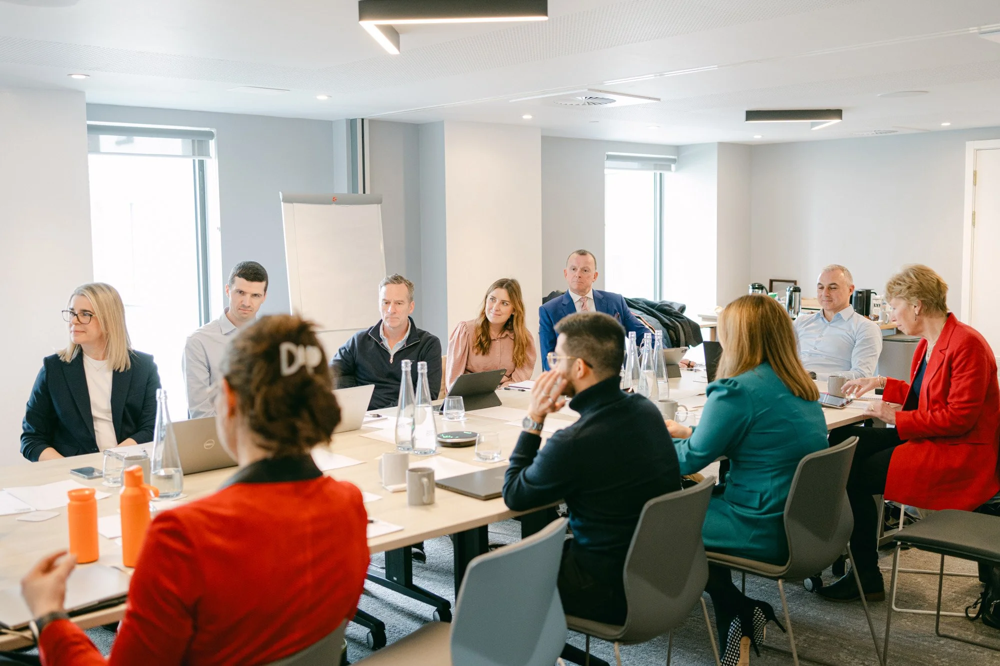 Business meeting with ten people seated around a conference table in a modern office, some with laptops and notebooks, engaged in discussion.