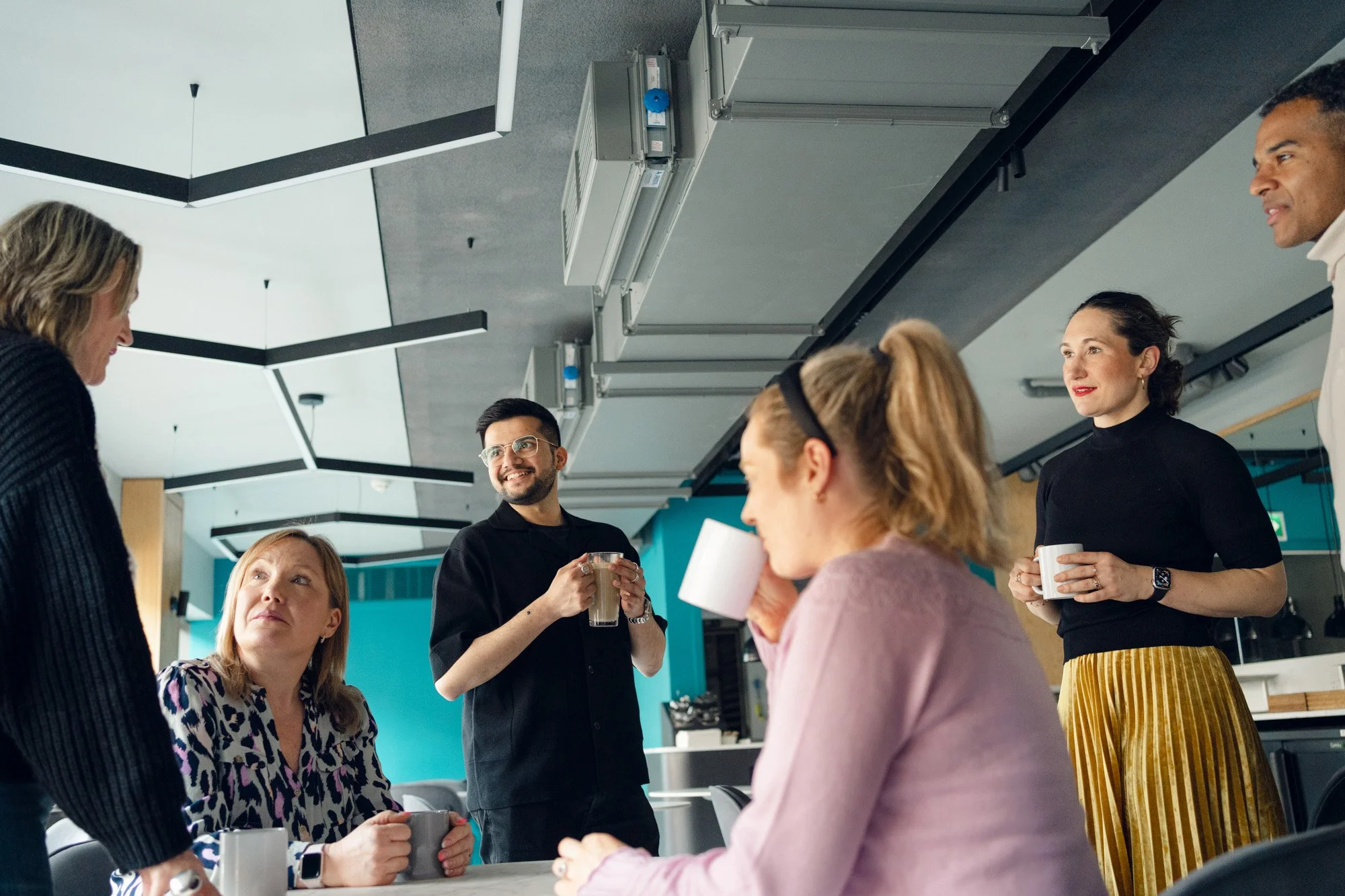 A group of six diverse coworkers in a modern office break room, with some seated at a table and some standing, engaged in conversation while holding coffee mugs.