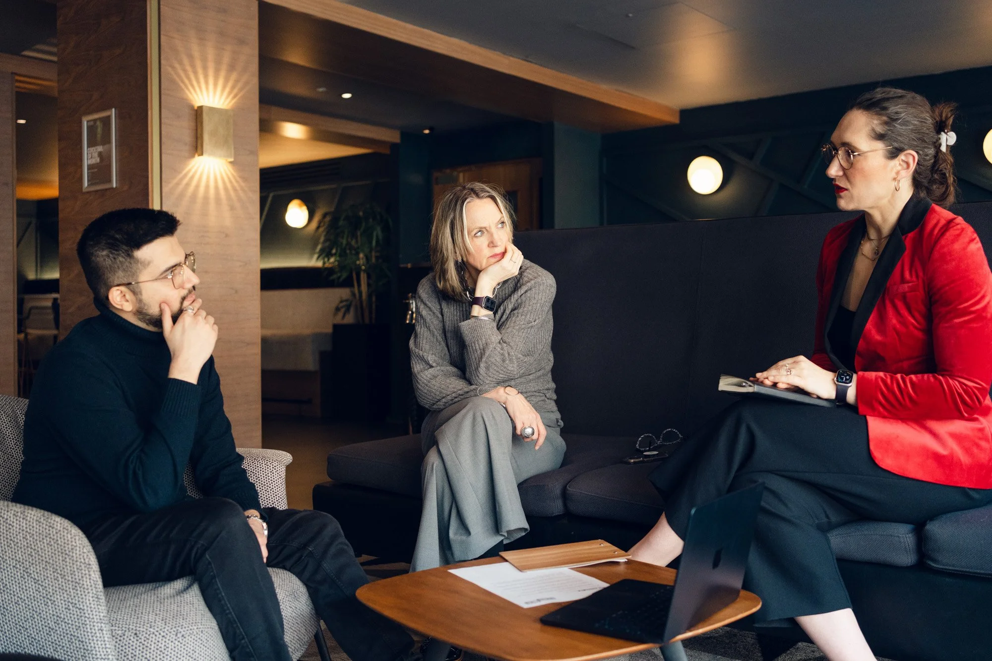 Three people having a discussion in a lounge area, with a laptop, papers, and glasses on the table.