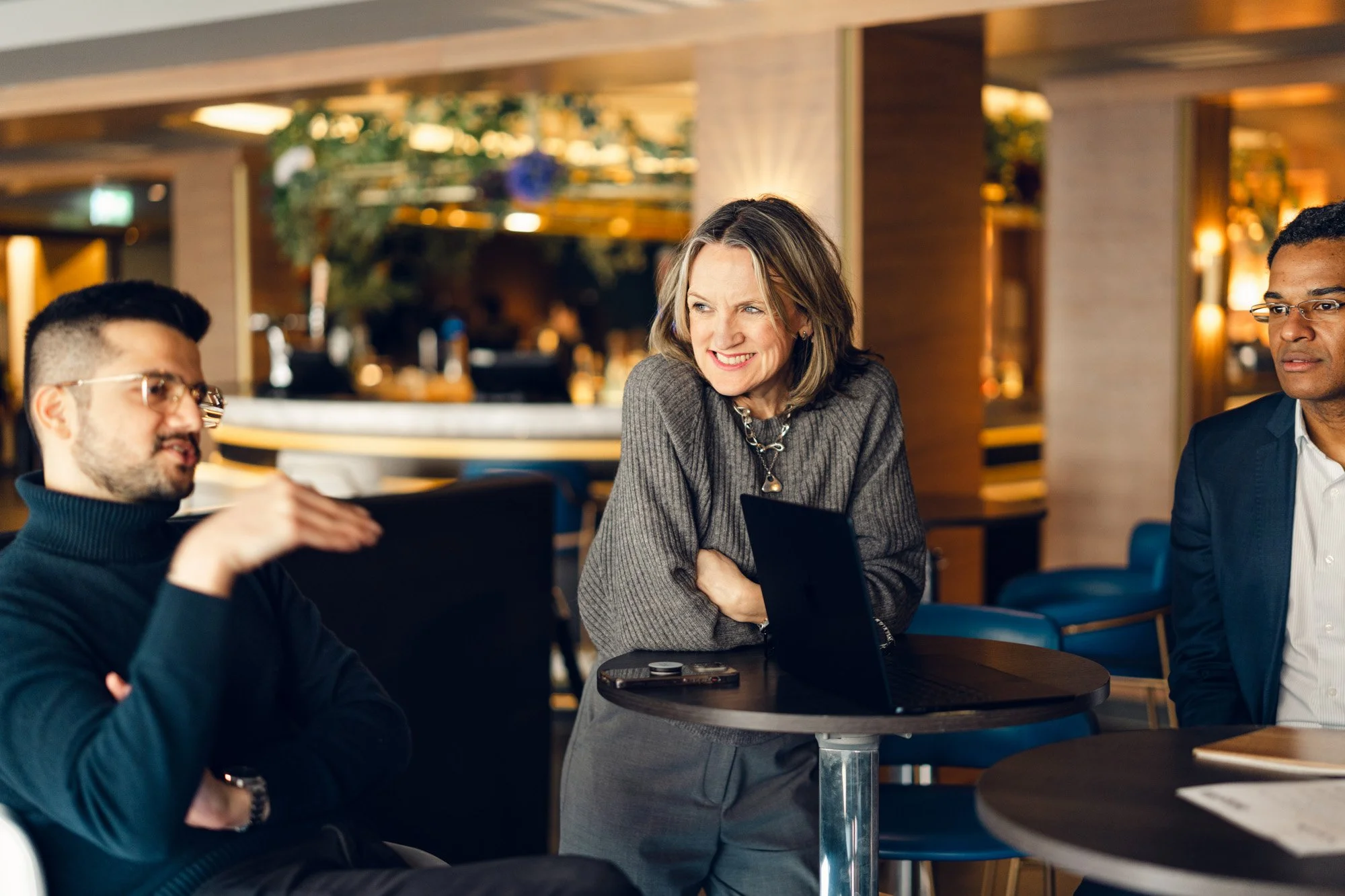 Three people having a conversation in a modern cafe, with a woman smiling and leaning on a small round table.
