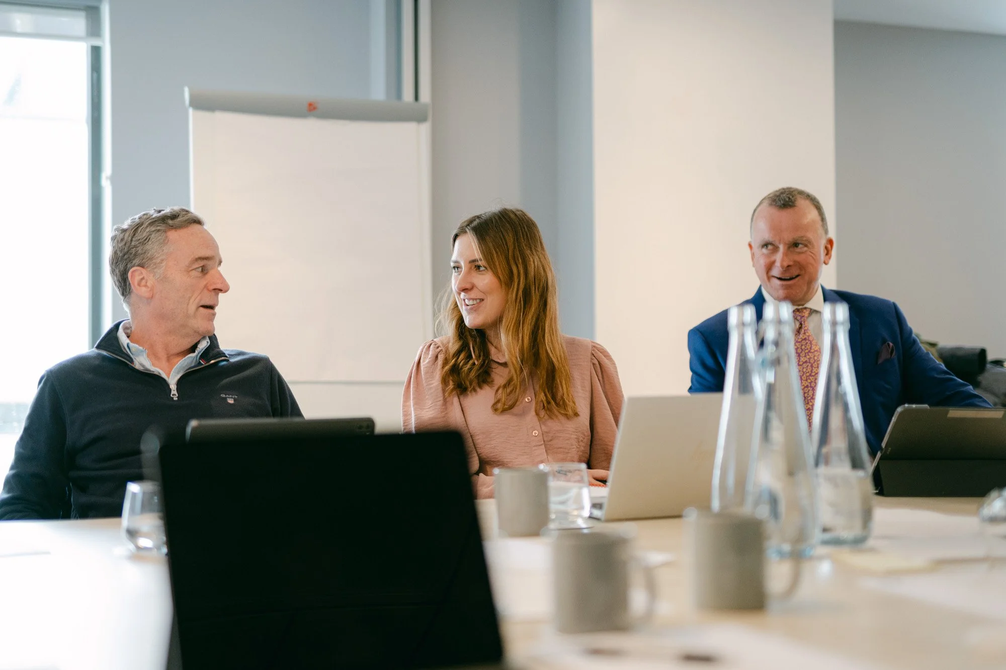 Three business professionals sitting at a conference table during a meeting, with laptops, glasses, and water bottles in front of them, engaging in conversation.