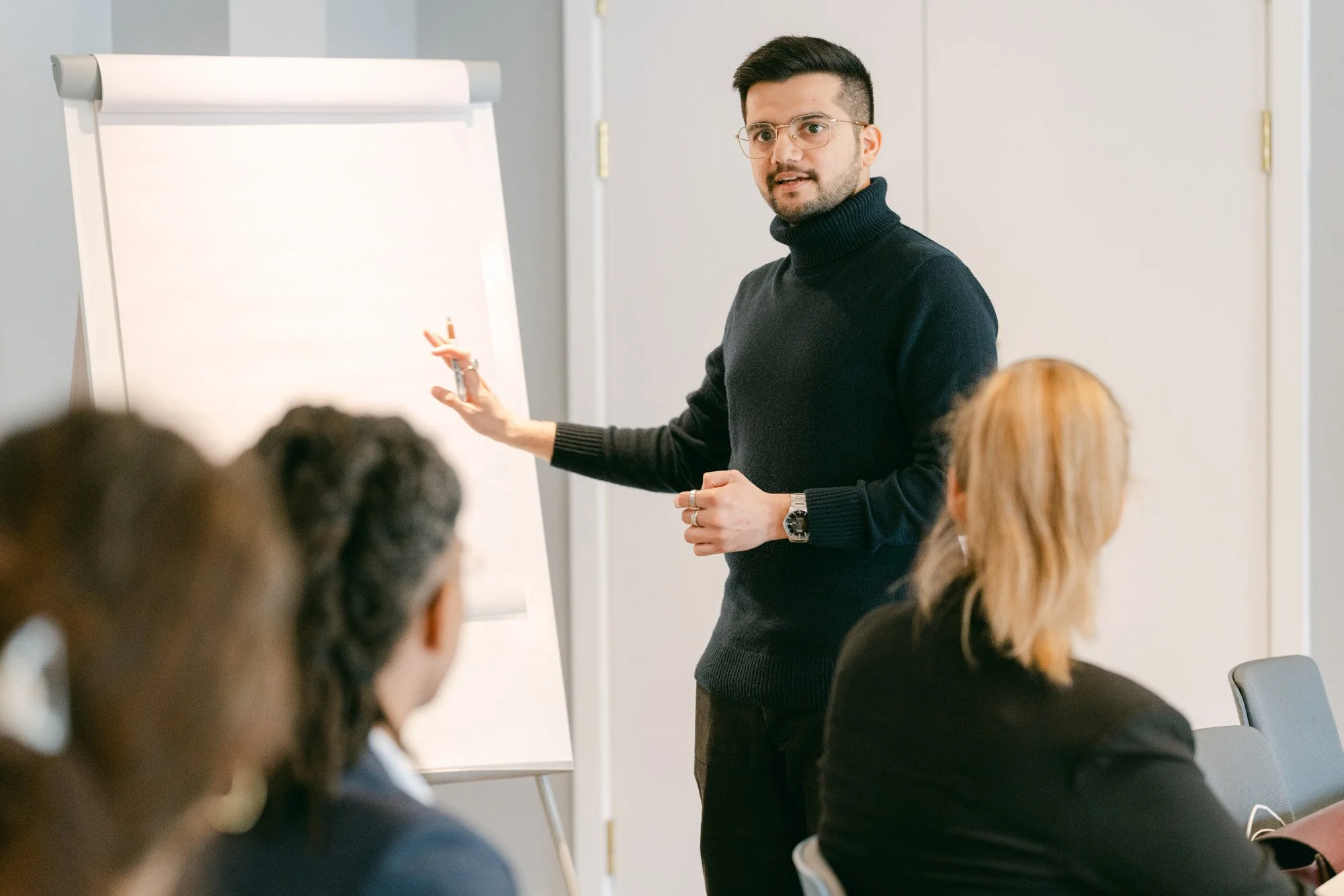A man with glasses and a black turtleneck sweater gives a presentation at a flip chart in a conference room. Several women are seated and listening.