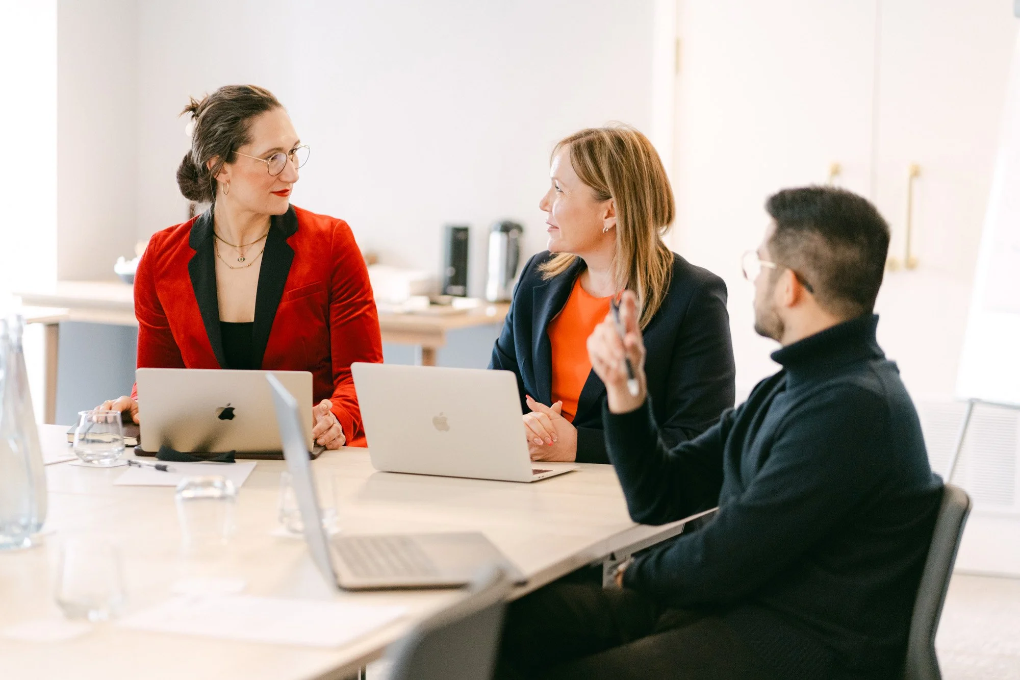 Three business professionals sitting at a conference table with laptops, engaging in a conversation in a bright office space.