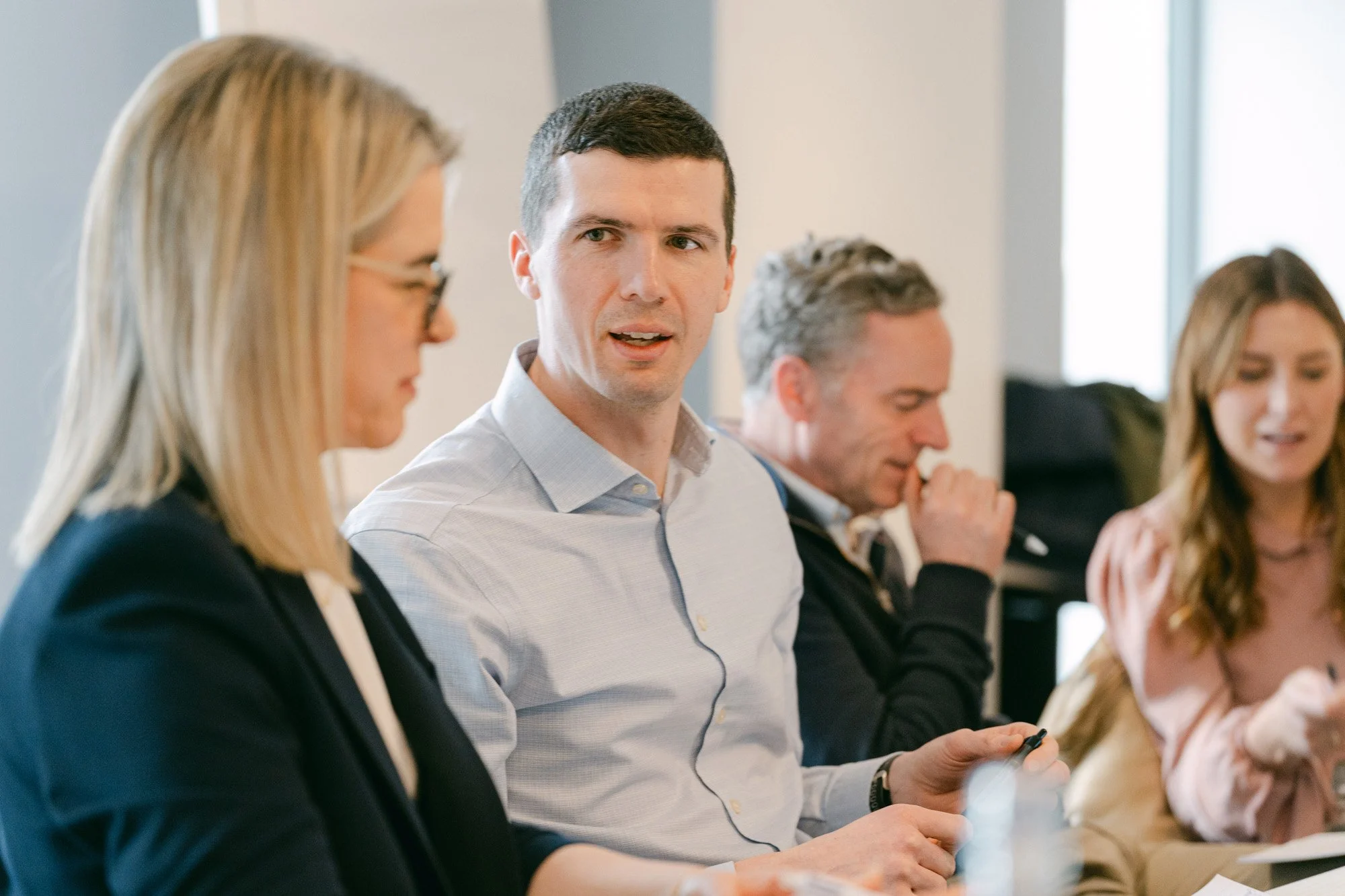 A group of people engaged in a discussion at a meeting or conference around a table, with one man speaking or listening, and others looking thoughtful or focused.