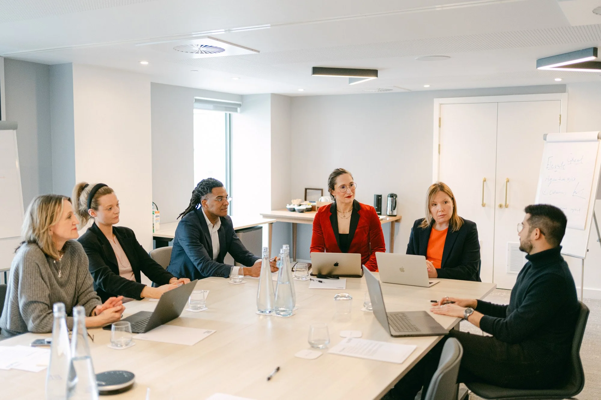 Business meeting with six people seated around a conference table, two laptops open, in a bright office, with a flip chart on a stand in the background.
