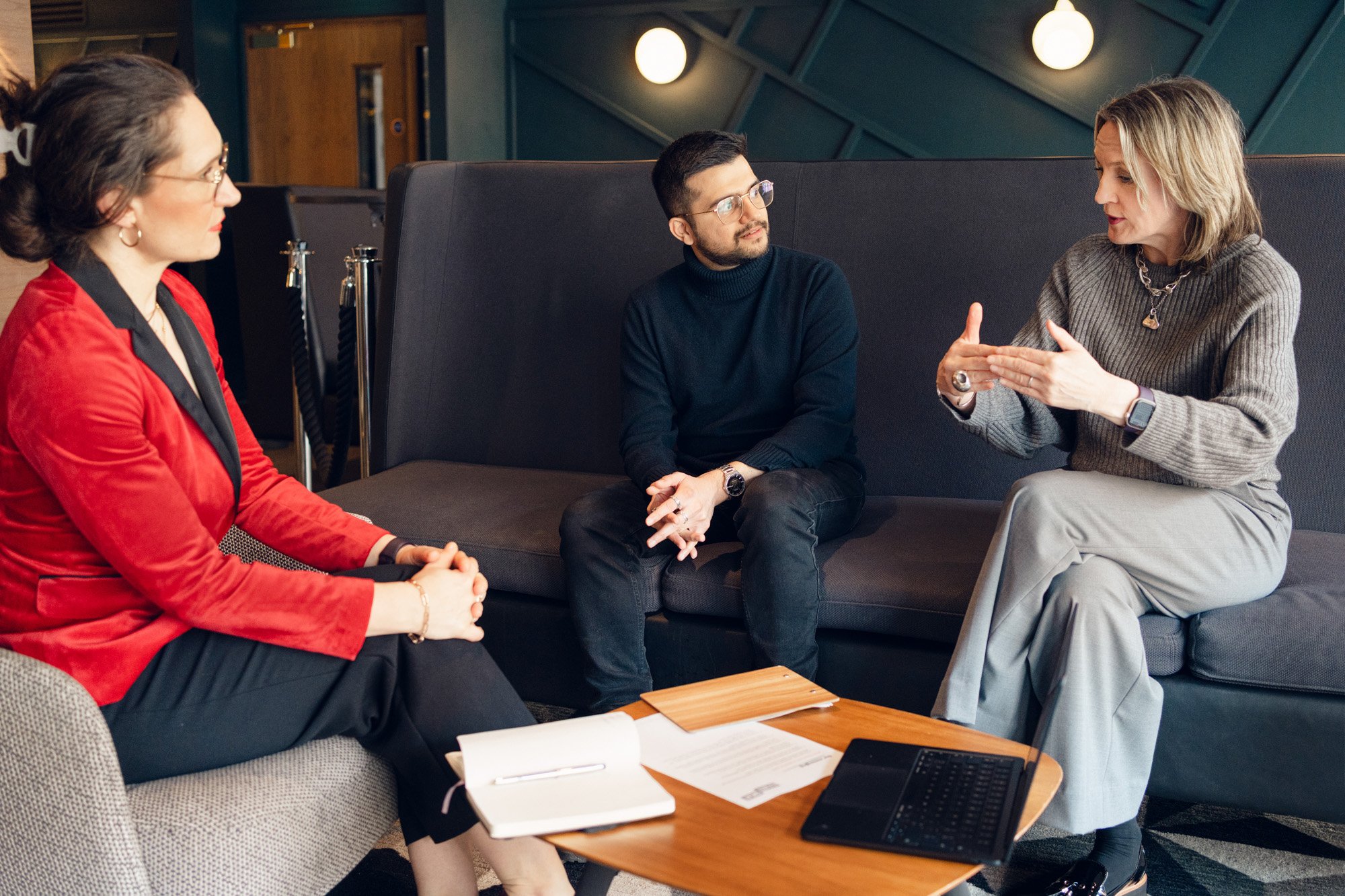 Three people having a discussion in a professional setting, two women and one man, sitting on couches and a chair around a small table with documents and a laptop.