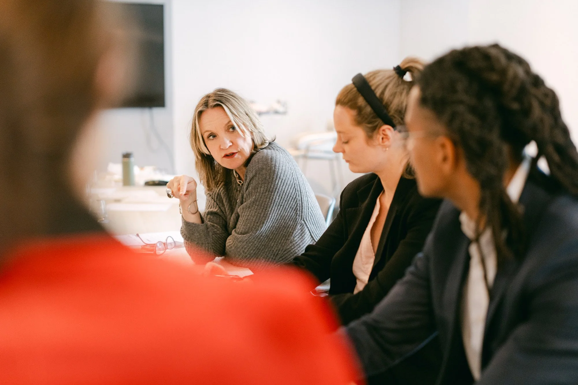A woman in a gray sweater is speaking and gesturing with her hand during a meeting, with three other women listening attentively, in a bright conference room.
