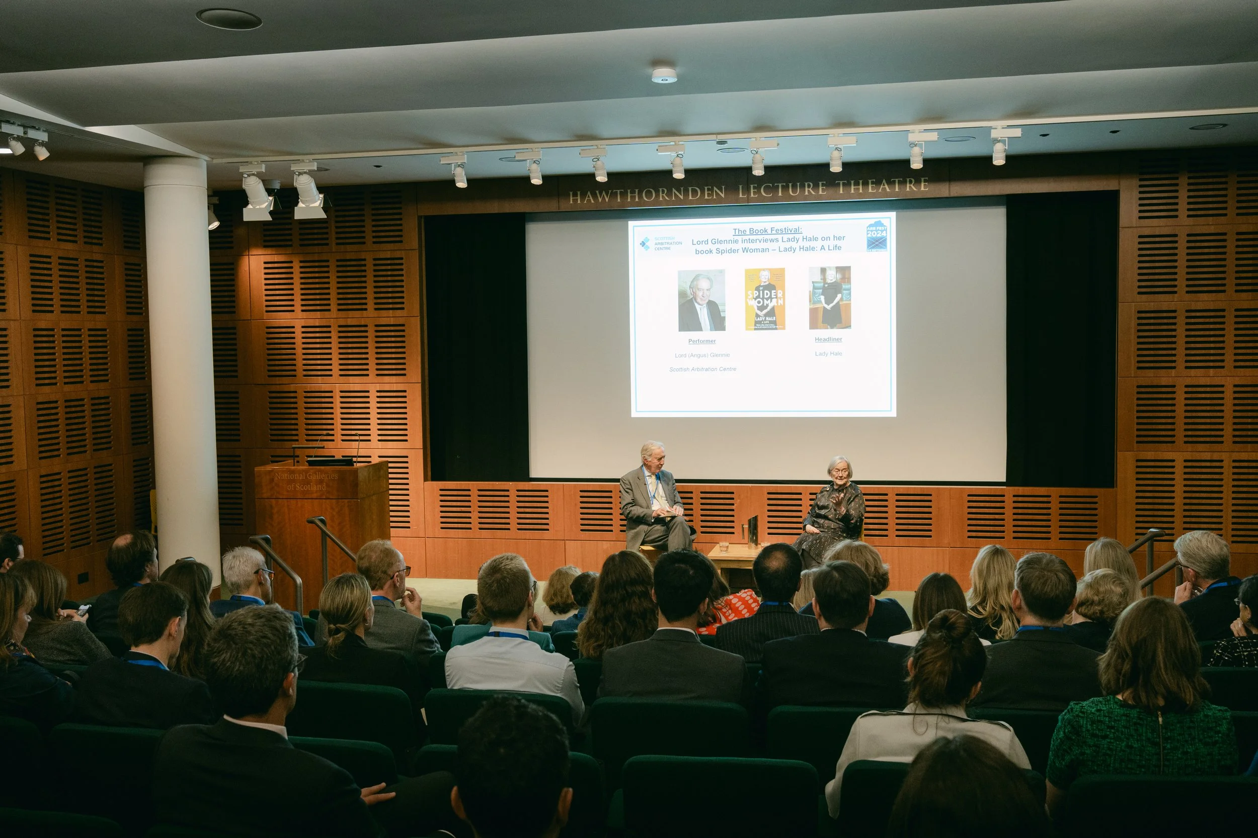 A lecture hall with an audience seated and two speakers on stage. The background features a large screen displaying information about a book festival and interviews with Lady Hale and Lord Glennie. The hall has wooden paneling and a sign that reads '