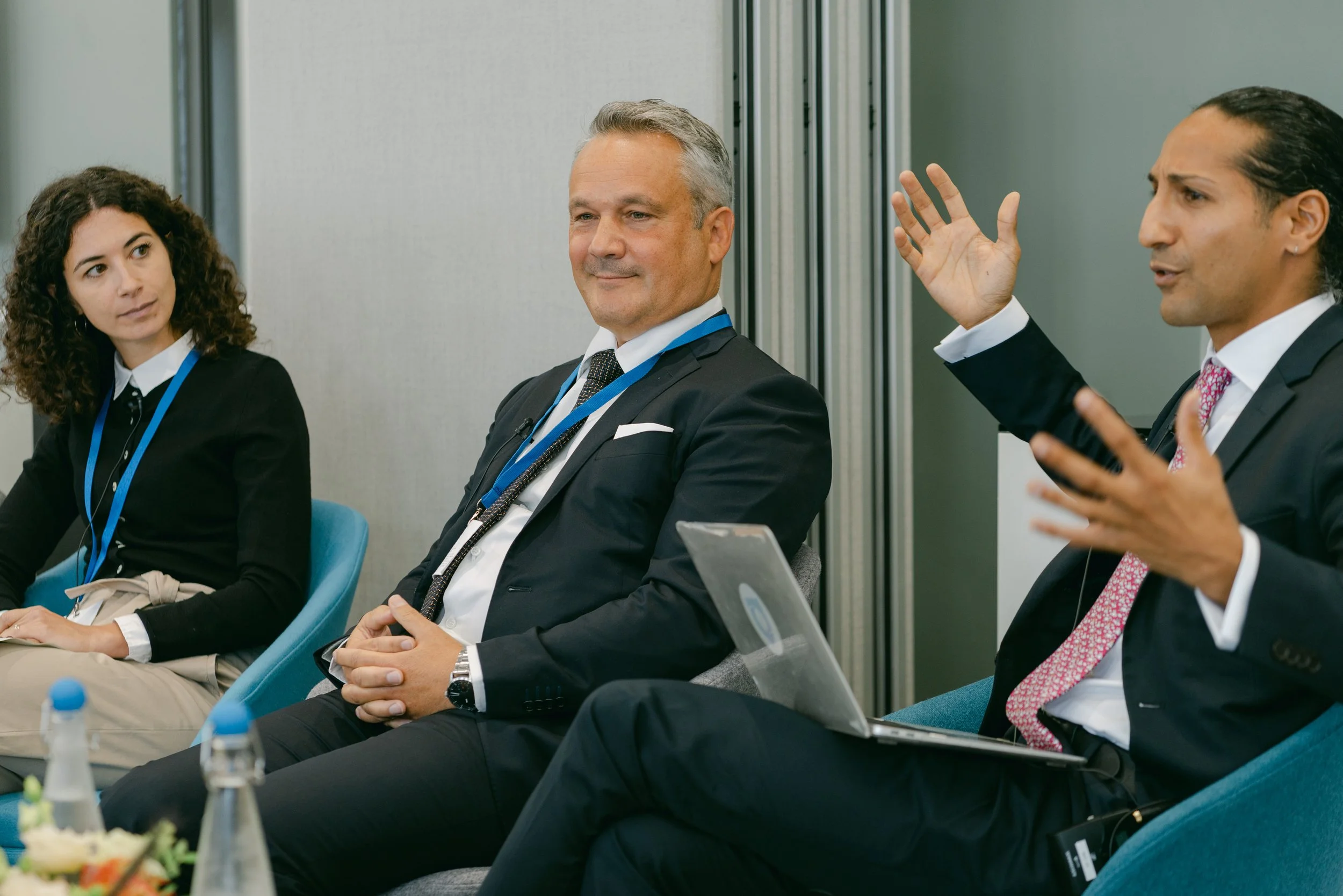 Three professionals in a discussion at a modern conference or meeting room, one man gesturing as he speaks, others listening, with laptops and bottled water on the table.