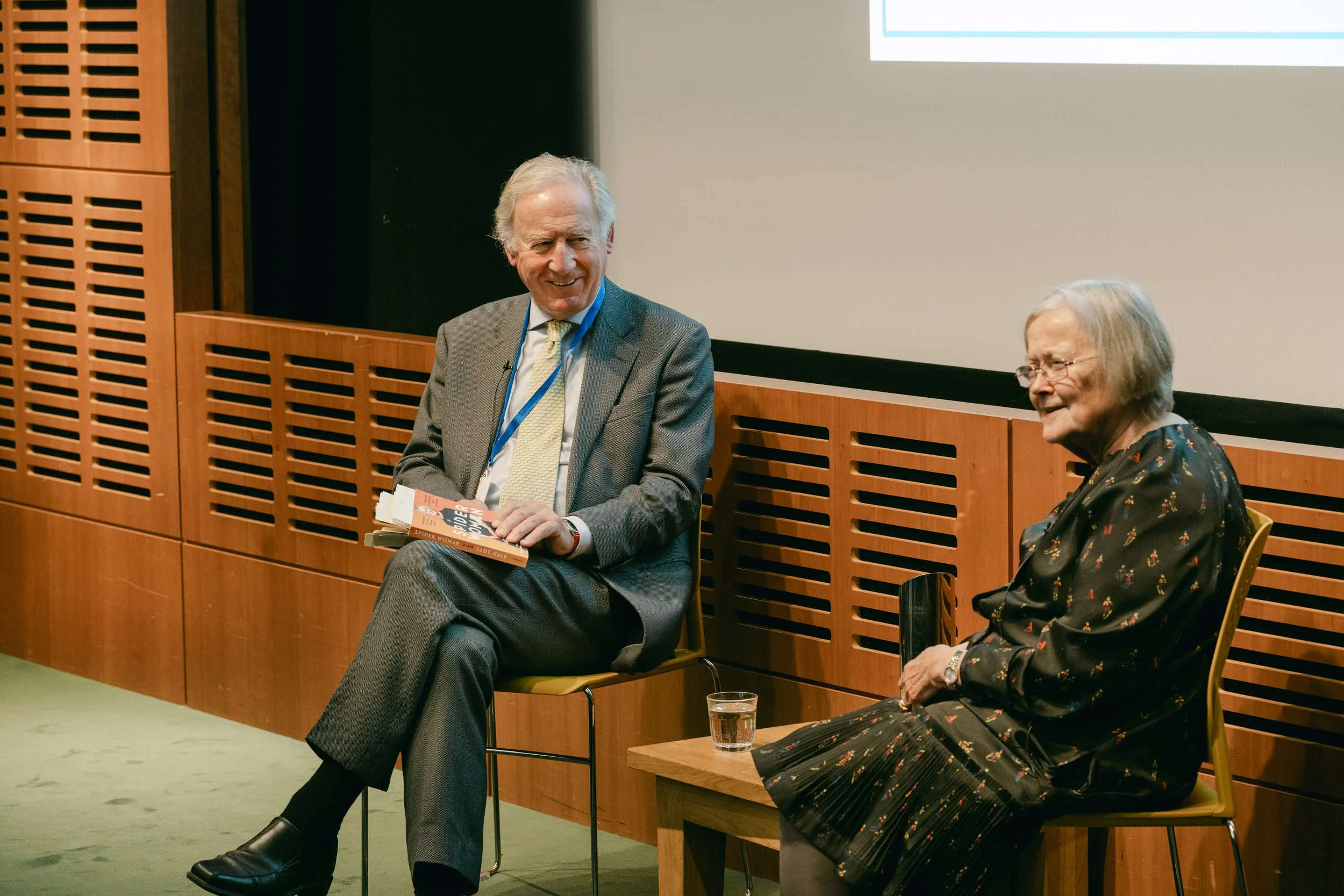 A man and a woman sitting and talking on stage during a panel discussion or interview.