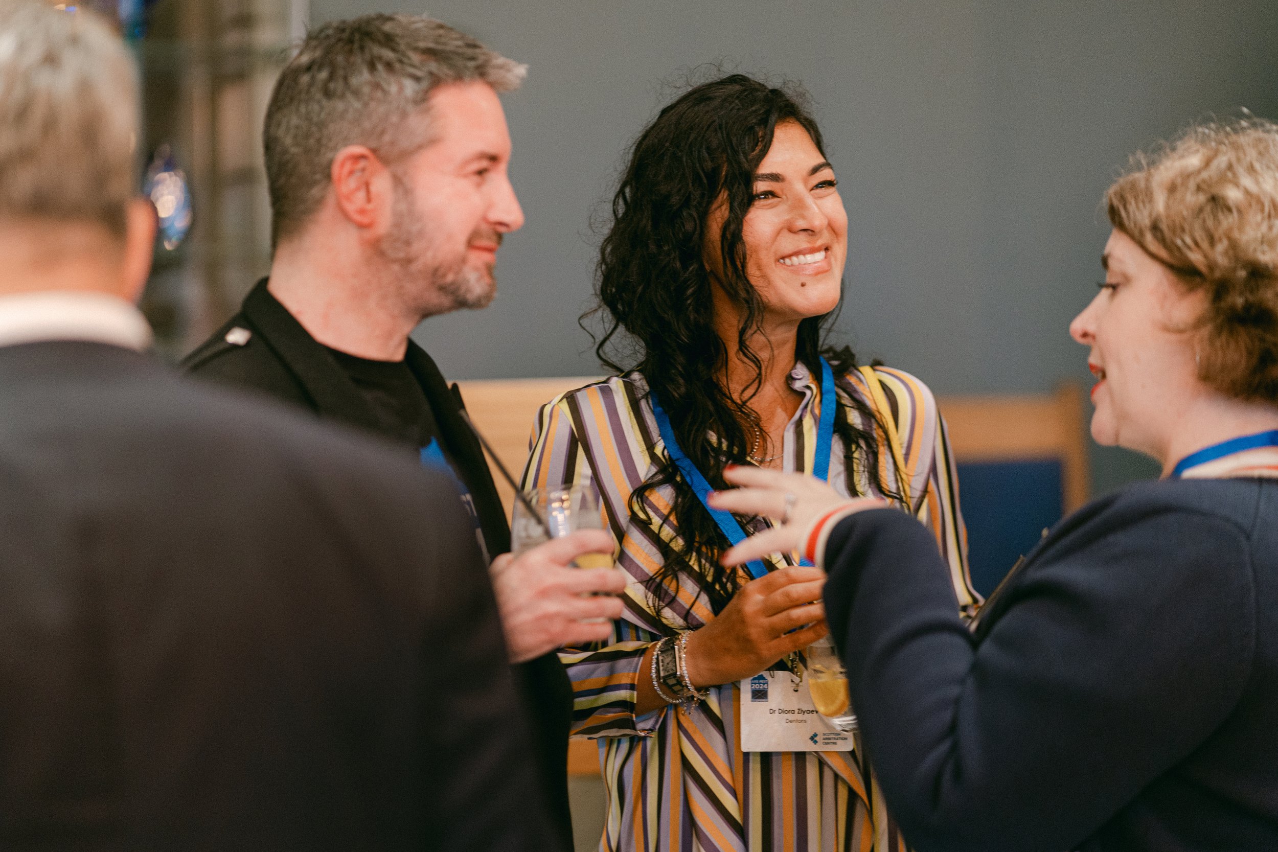 Group of four people talking and smiling at a networking event, one woman with curly black hair, wearing a striped dress, holding a drink, and wearing a conference badge.