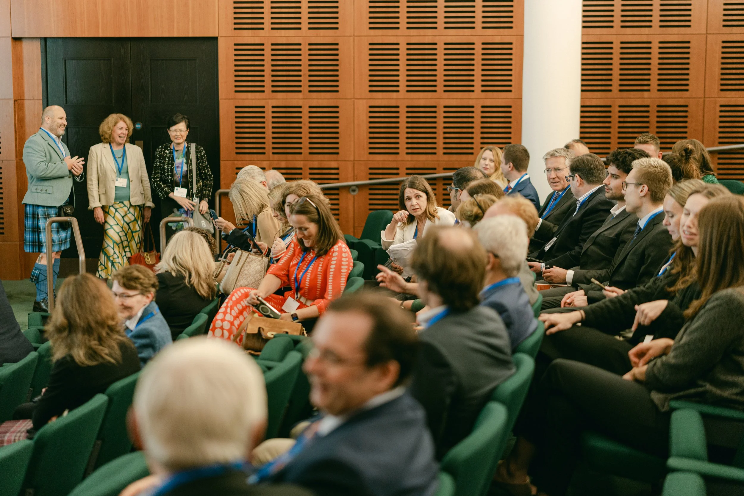A group of people seated in a conference room, some engaging in conversation, while three individuals stand near the back, smiling and talking.