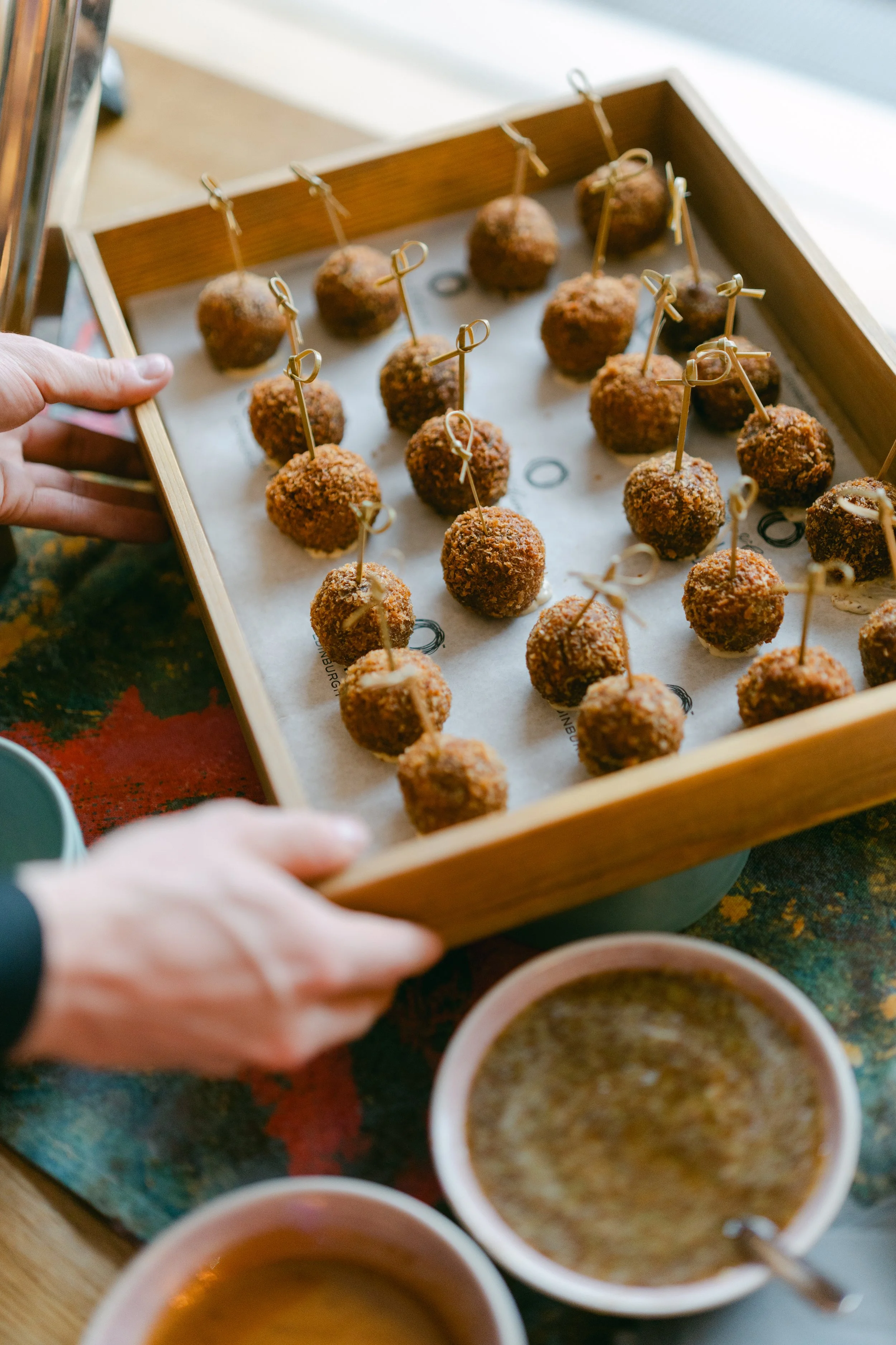 Tray of fried appetizers, possibly croquettes or arancini, with skewers, on a table with small bowls of sauce nearby.