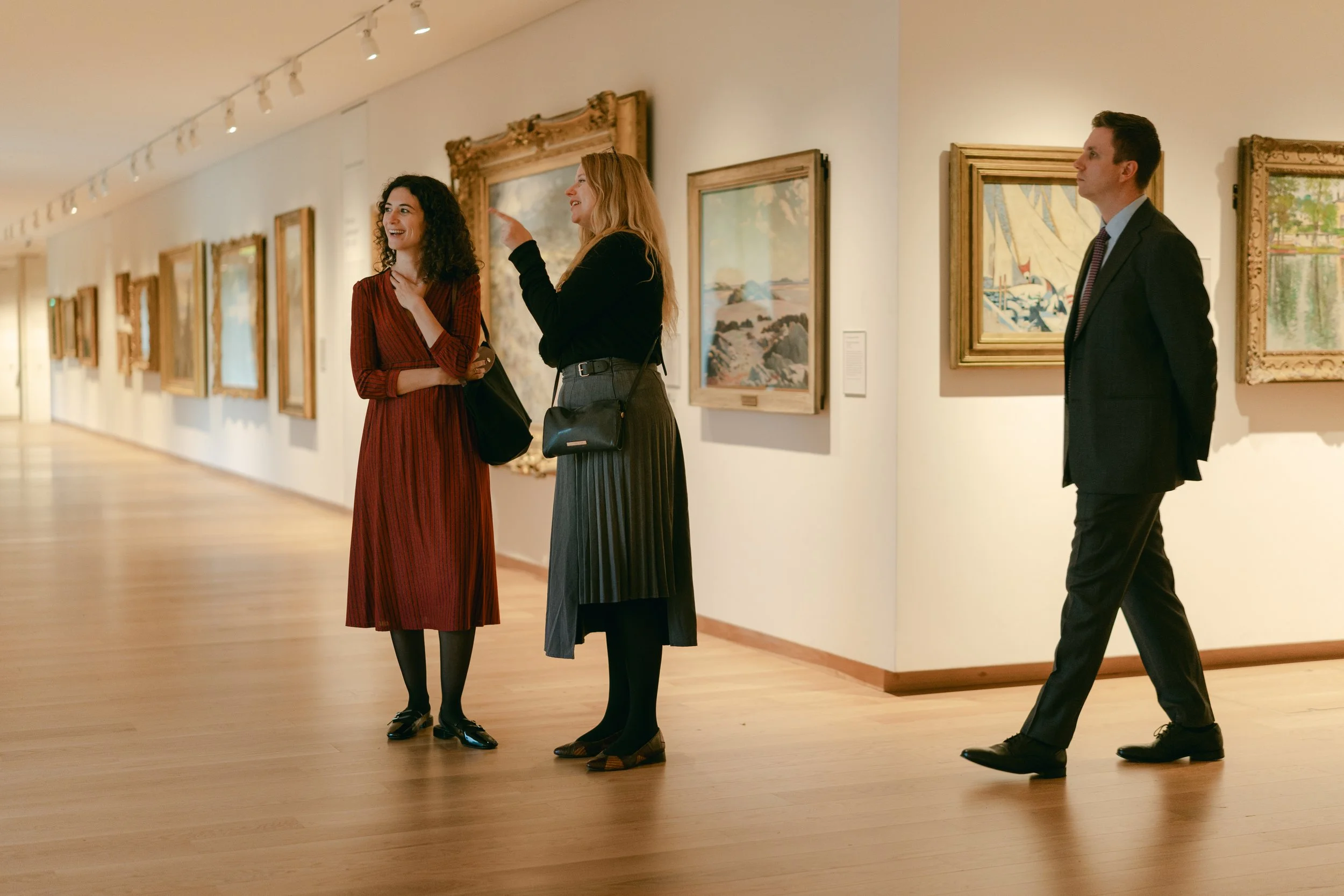 Three people in an art gallery. Two women are talking and smiling, one wearing a red dress and the other a black top and gray skirt. A man in a suit walks past them, looking at the paintings on the wall.