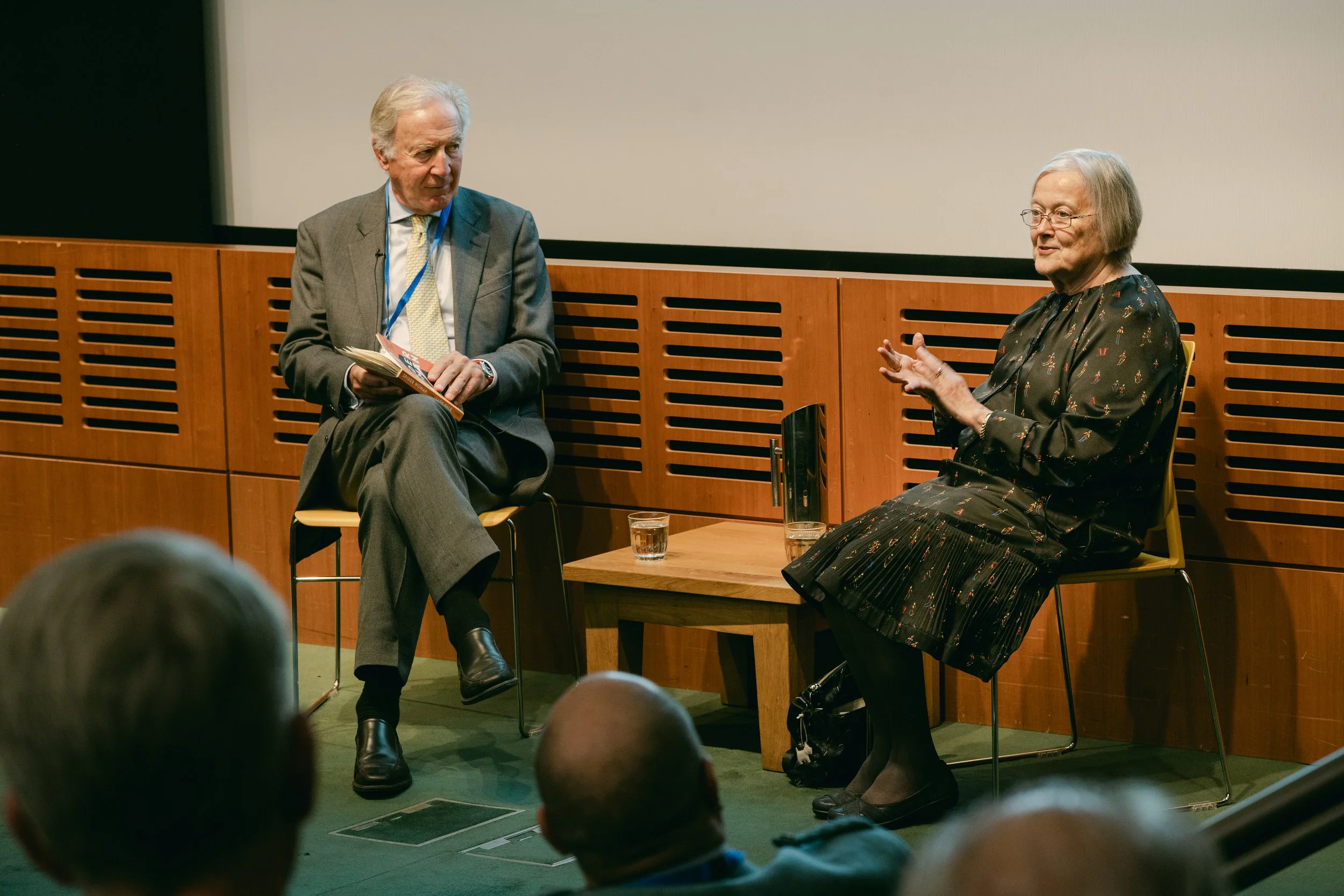An elderly man and woman seated on stage in conversation, with an audience visible in the foreground.