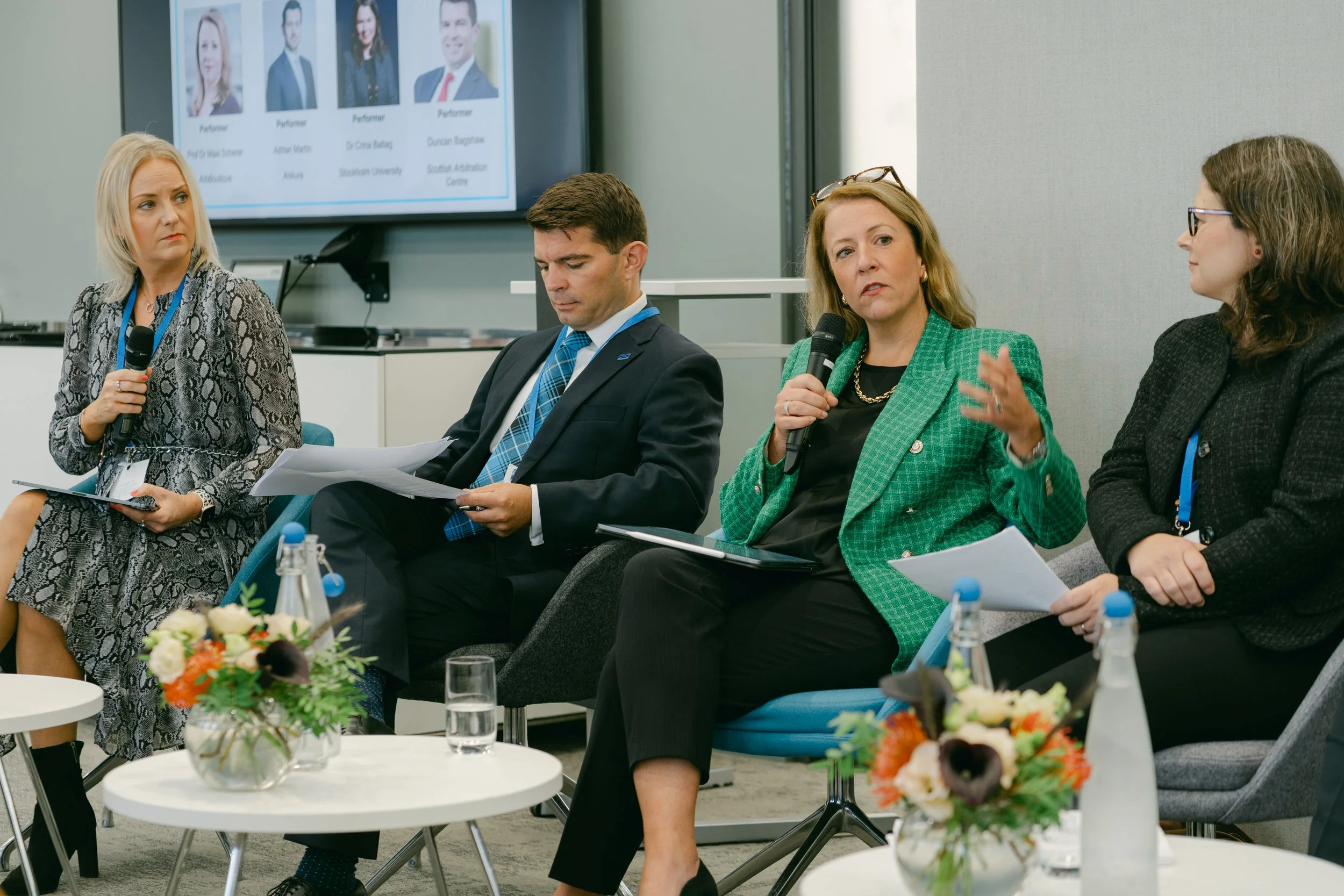 Four people participating in a panel discussion at a professional conference. A woman in a green blazer is speaking into a microphone. Others are listening, with one woman holding a microphone and two others looking at papers. There is a screen in th