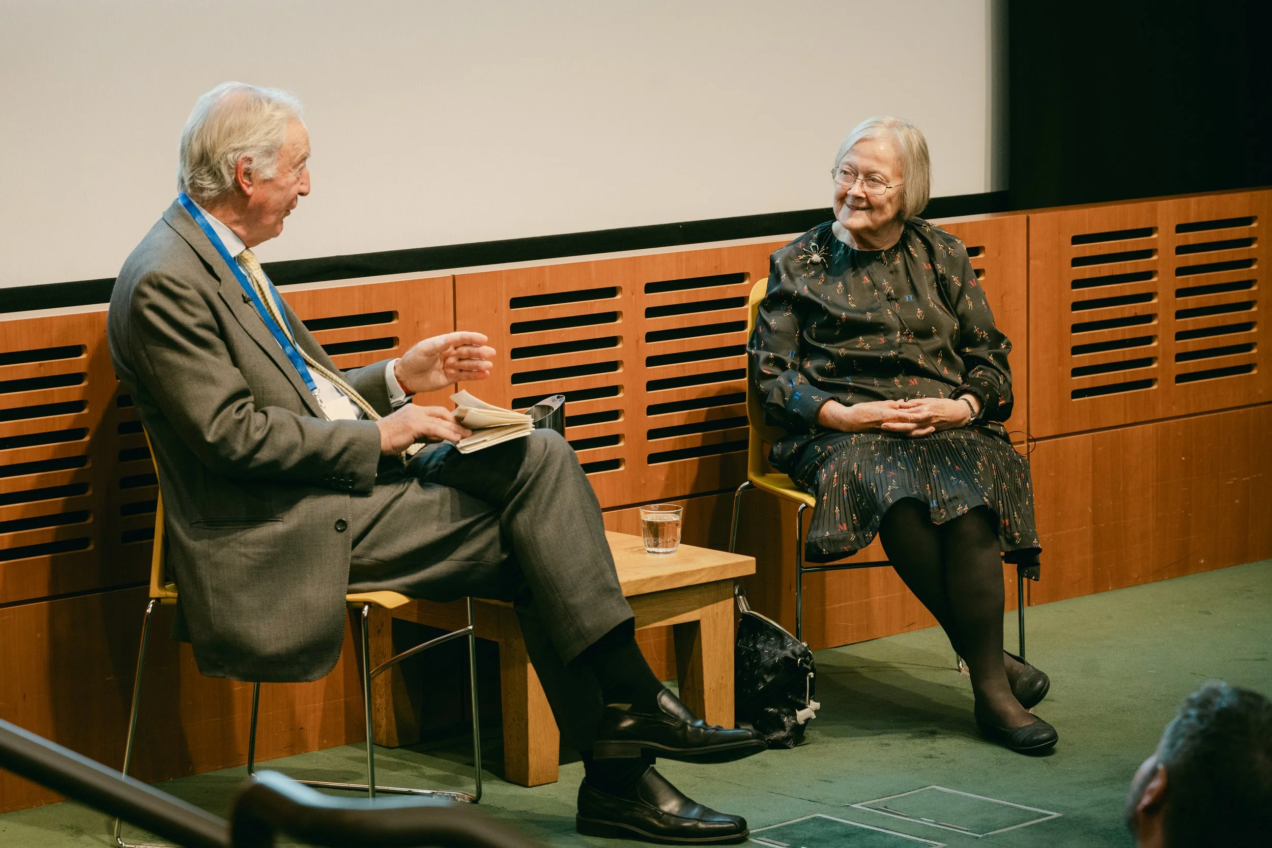 An older man and woman are seated on stage in conversation, with the man holding a notebook and the woman smiling and listening, in front of a large white screen and wooden paneling.