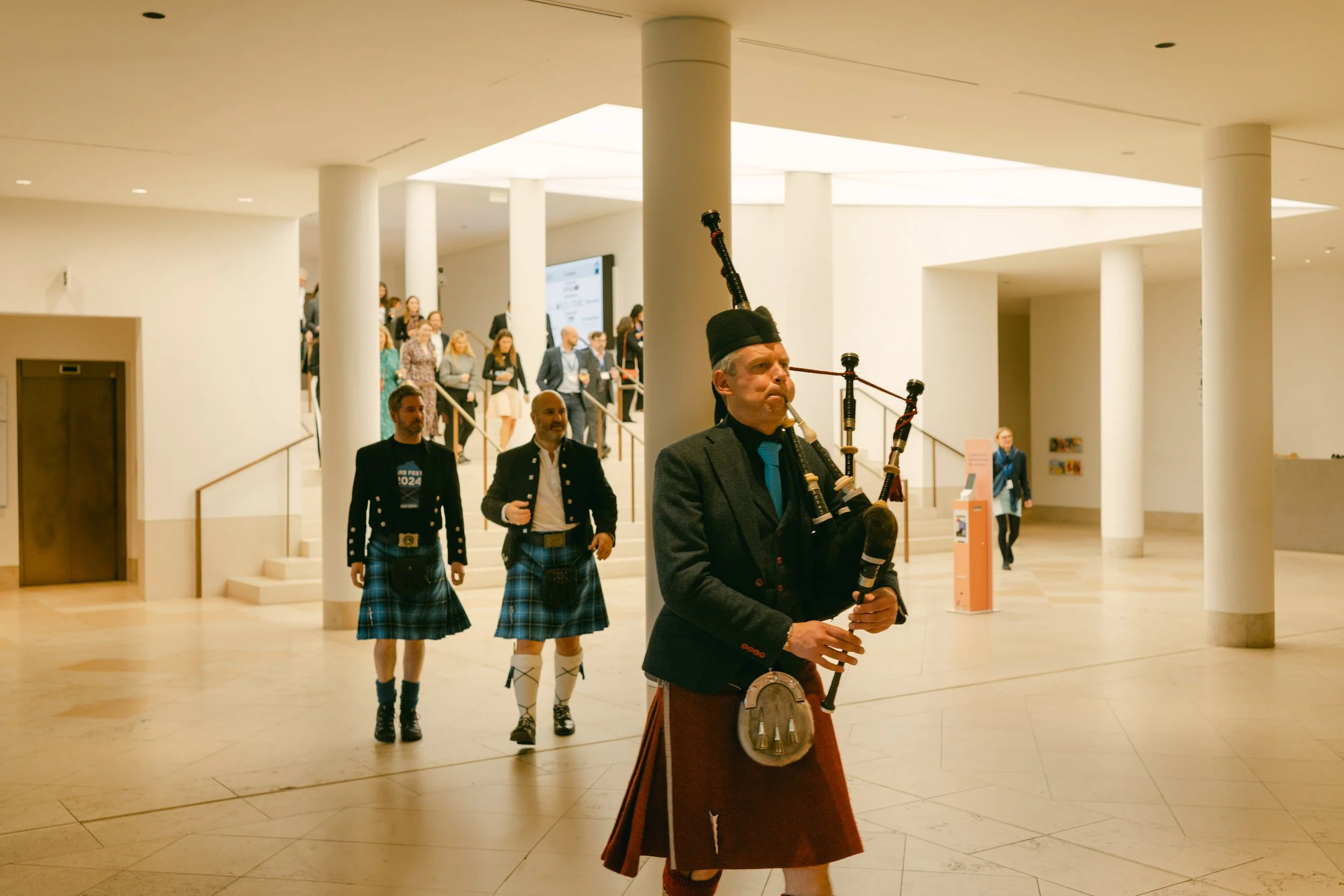 A man playing bagpipes in an indoor public space, with a group of men in kilts walking in the background and a large group of people on stairs.