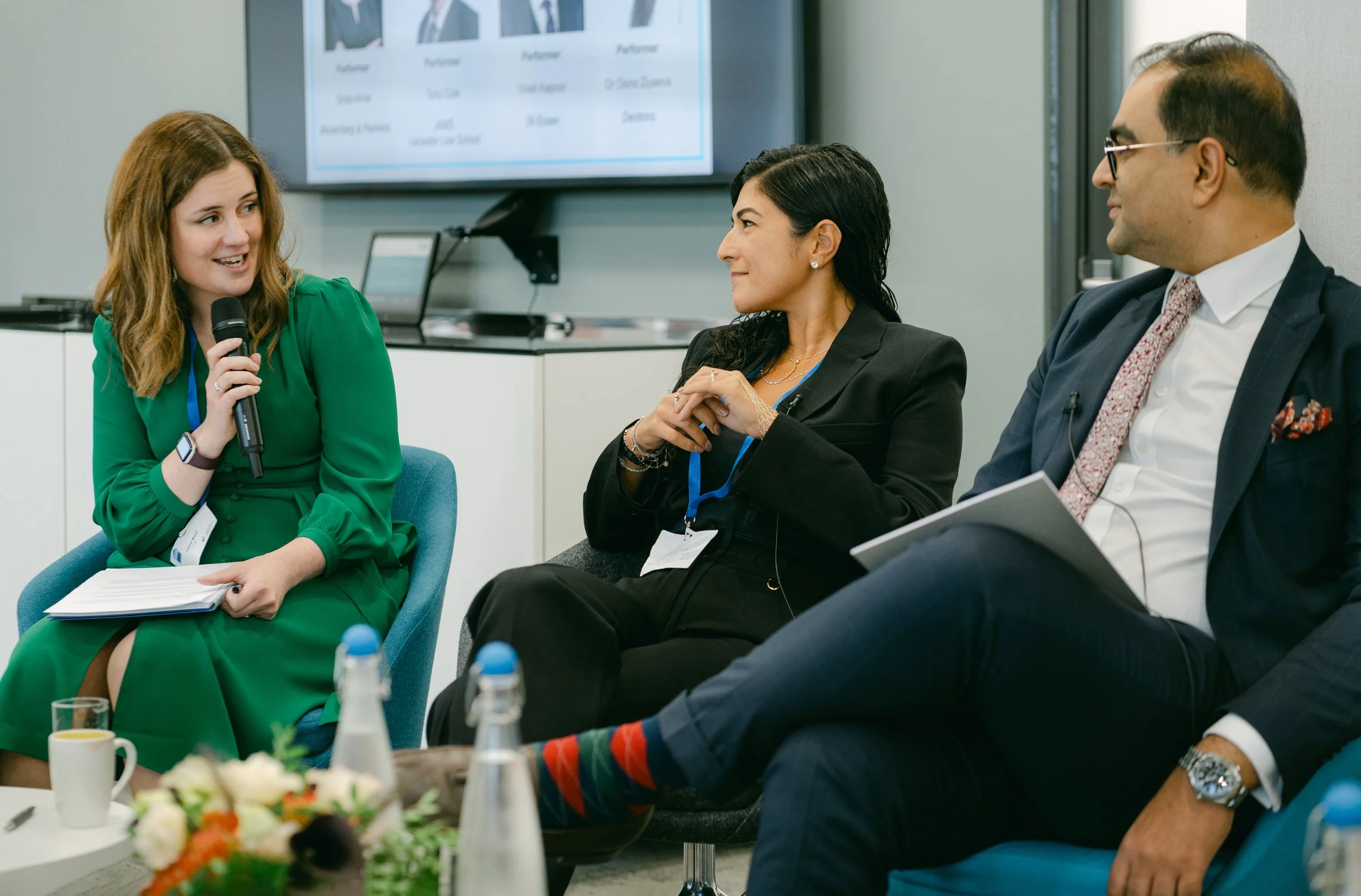 Three professionals engaged in a panel discussion in a conference room, with a woman in a green dress holding a microphone on the left, a woman in black business attire in the middle, and a man in a suit and tie on the right, facing each other.