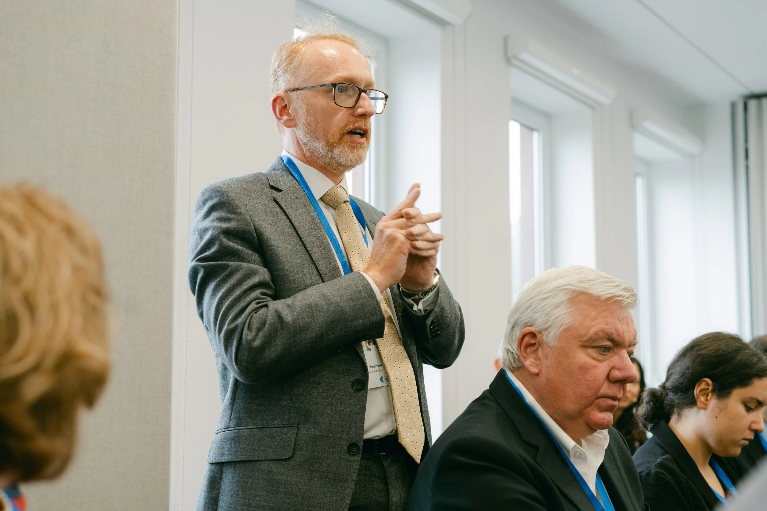 A middle-aged man in a gray suit, white shirt, and polka dot tie is speaking during a meeting or conference. He has glasses and is making a hand gesture. Seated at the table are other individuals, including an older man with white hair and a woman wi