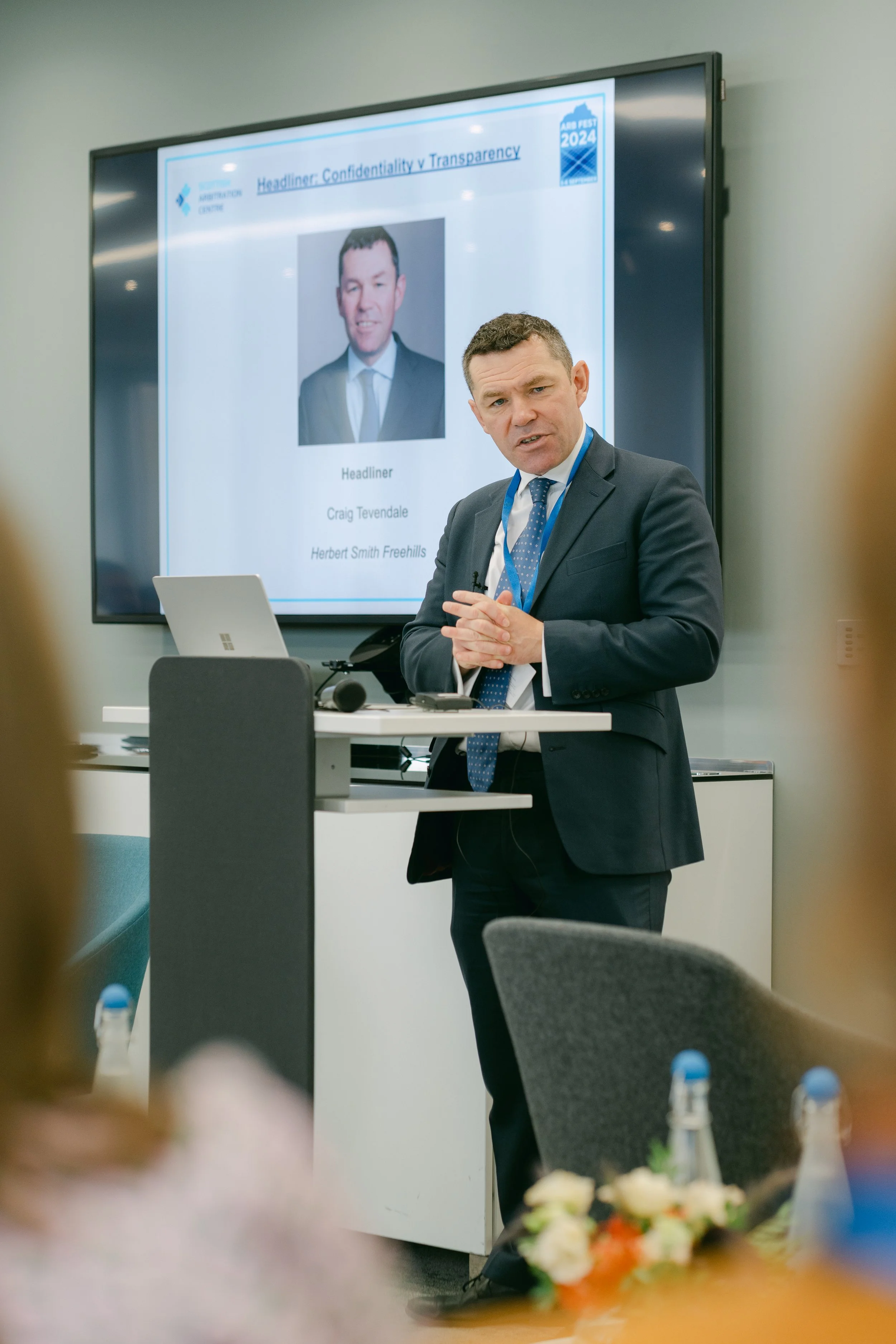 A man in a business suit giving a presentation in a conference room, with a large screen behind him displaying a slide titled 'Headliner Confidentiality v Transparency' and a headshot of a man named Craig Tevendale, alongside the names Herbert Smith 
