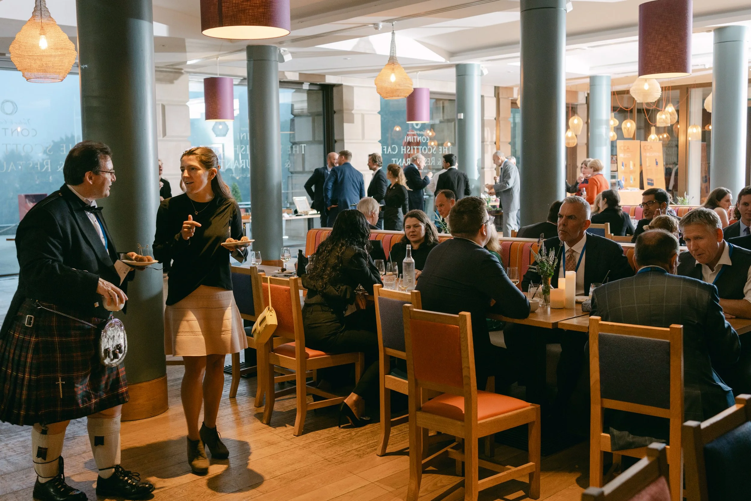 People enjoying a formal meal and networking at a restaurant or event space, with waitstaff serving food in the foreground and guests seated at tables in the background.
