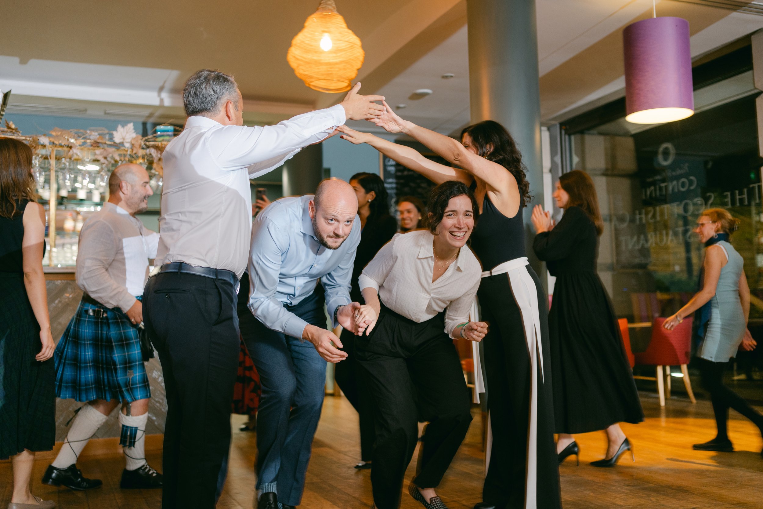 People dancing and celebrating in a festive atmosphere at a social gathering or party, with some holding hands and others clapping.