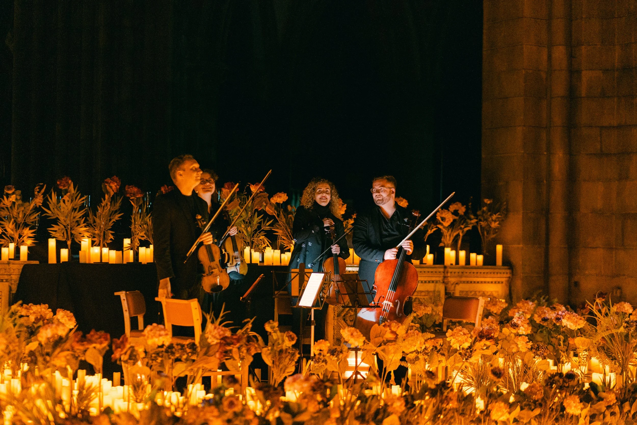 A group of musicians performing on stage decorated with candles, flowers, and foliage, holding string instruments in a dimly lit setting.