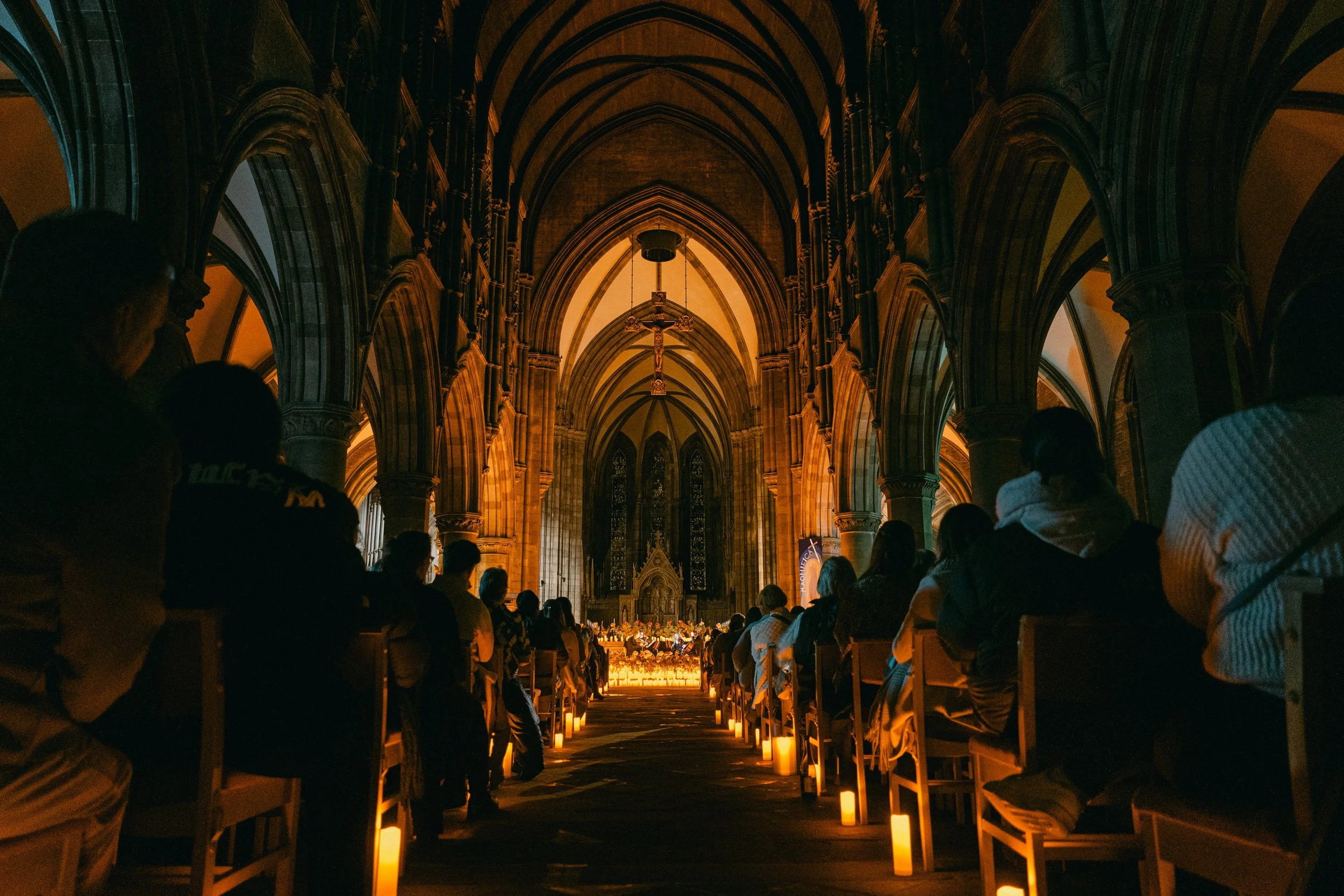 People attending a religious service inside a grand cathedral with high vaulted ceilings, stained glass windows, and candles along the aisle.