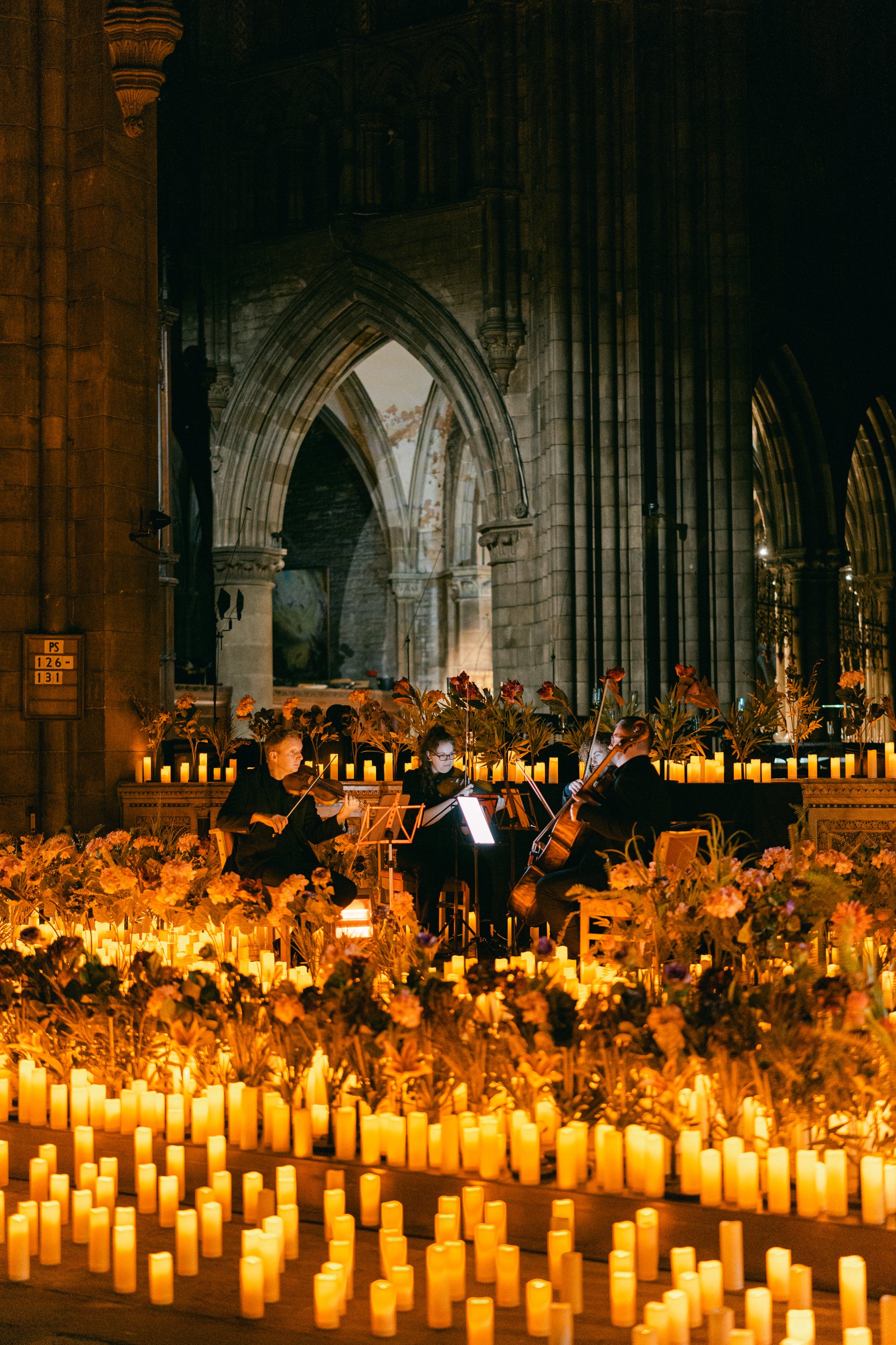 Three musicians playing string instruments in a dimly lit, candle-lit church setting with gothic arches and floral arrangements.