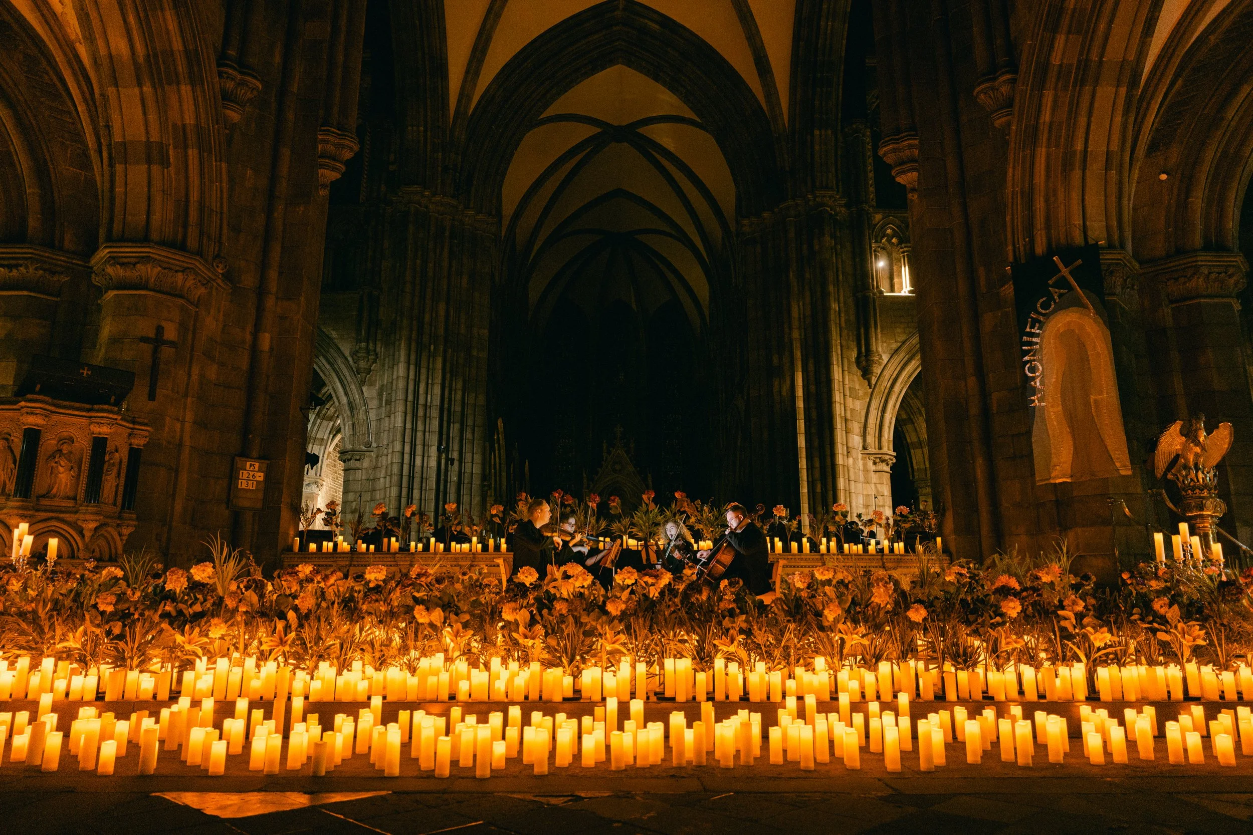 Inside a large Gothic-style church with a high arched ceiling, lit by numerous candles, with a few musicians playing string instruments surrounded by flowers.