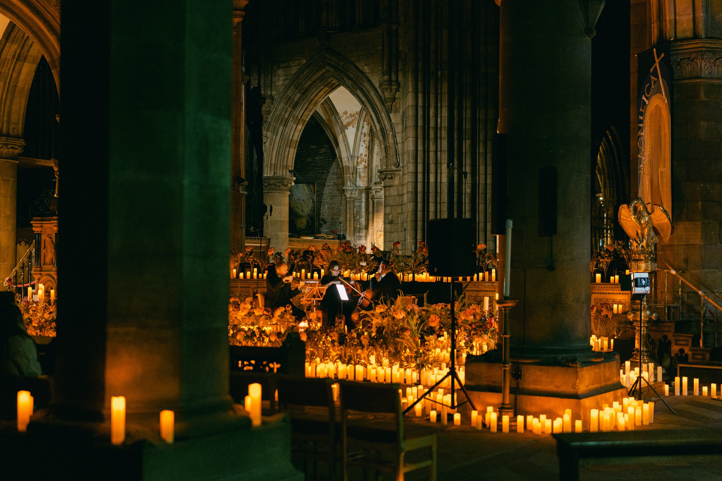 A musical performance inside a church, with musicians playing string instruments among many lit candles and flowers, illuminated by warm lighting, with church architecture visible in the background.