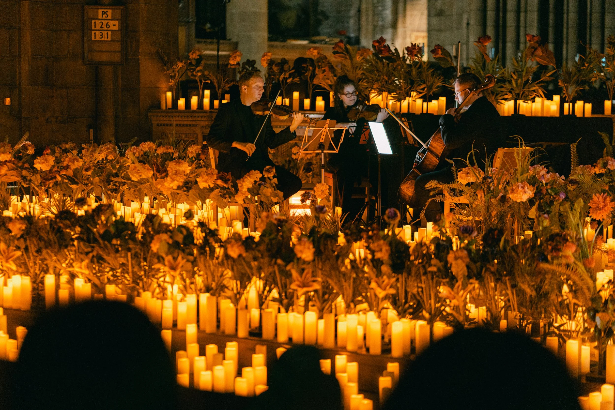 Three musicians playing string instruments on a stage decorated with numerous candles and flowers in a dimly lit indoor setting.